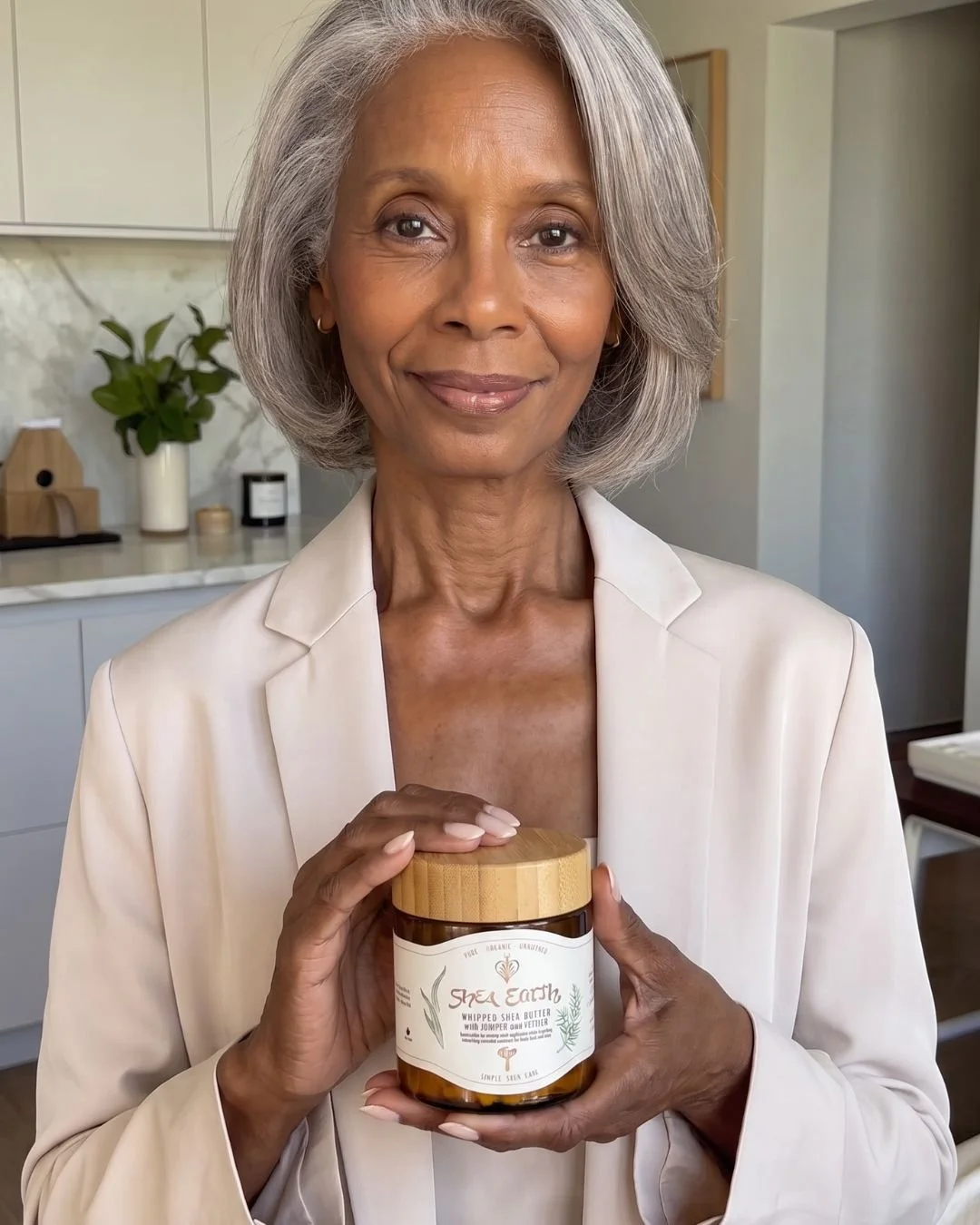 An elderly woman with gray hair holding a jar of Shea Earth whipped shea butter in a modern kitchen.