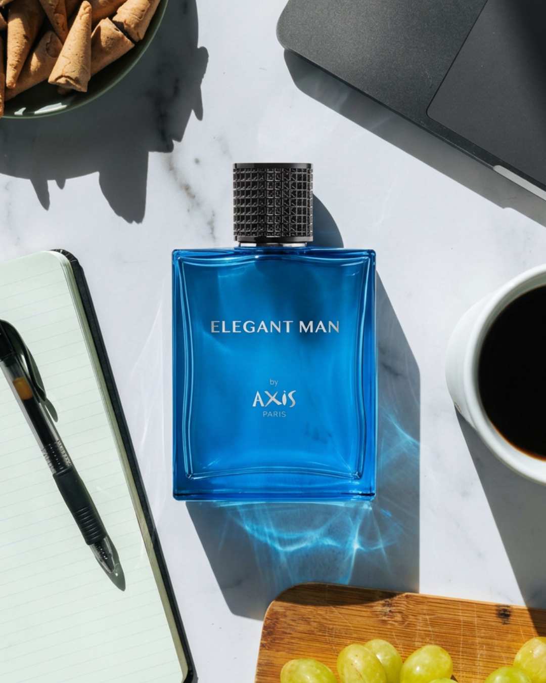 A blue bottle of men's perfume labeled "Elegant Man by Axis Paris" on a white surface, surrounded by a notebook with a pen, a bowl of rolled wafers, a cup of black coffee, a laptop, and a wooden tray with green grapes.