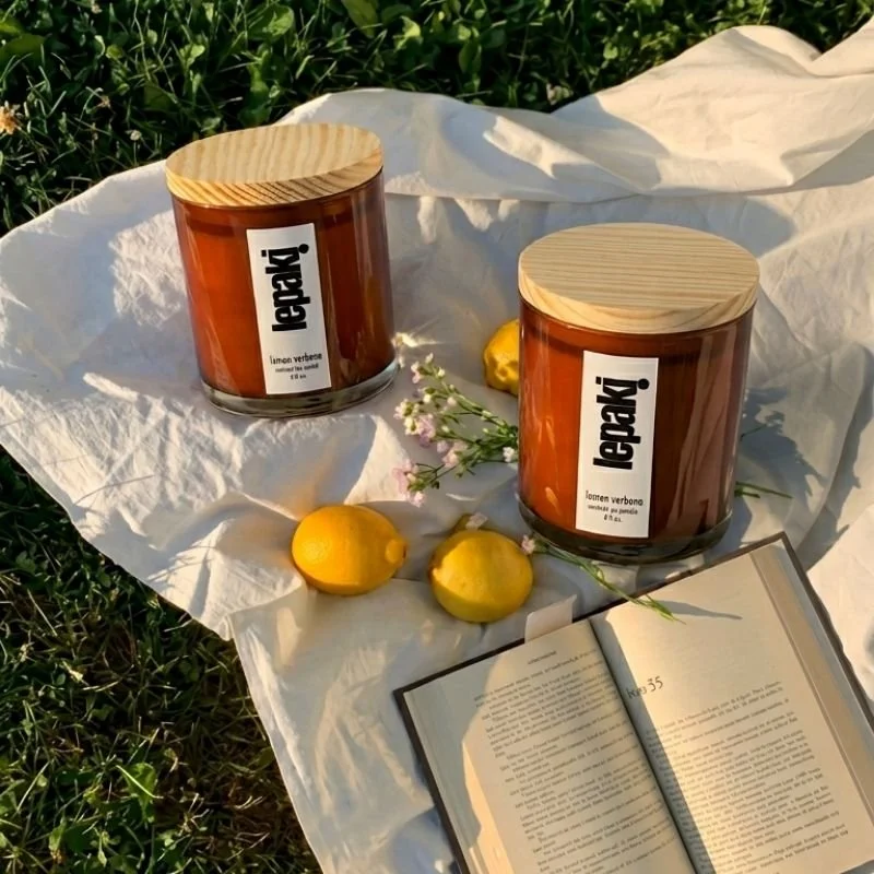 Two candles in brown glass jars with wooden lids, labeled in Spanish, placed on a white cloth with lemons, small pink flowers, and an open book.