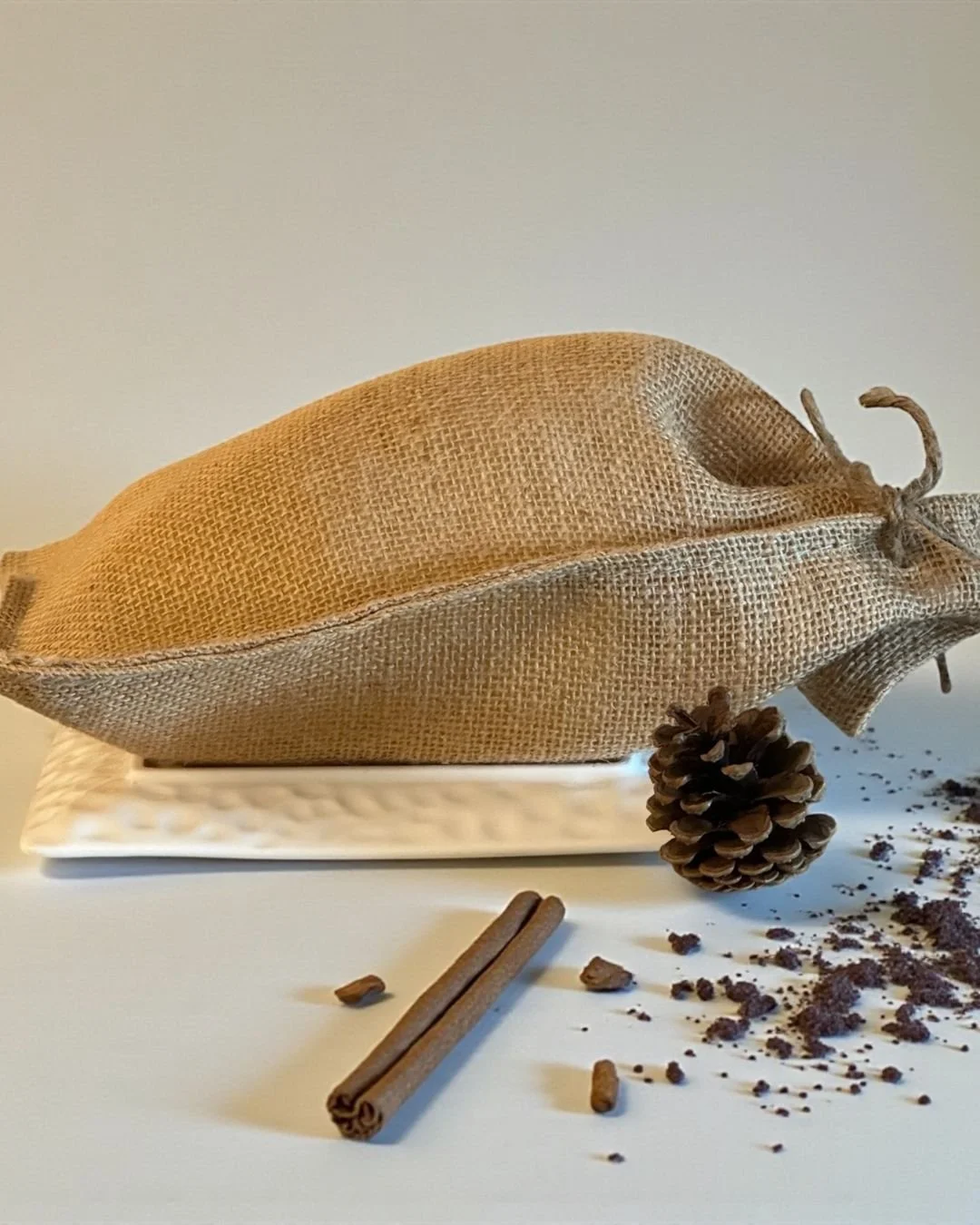 A burlap sack resting on a white rectangular tray, surrounded by pinecones, cinnamon sticks, and scattered coffee grounds on a white surface.