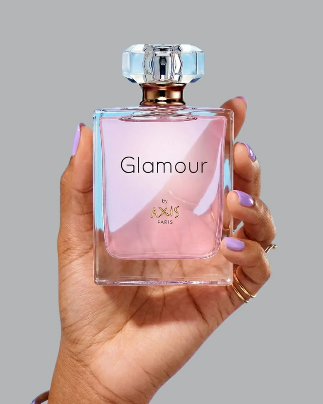 A hand holding a pink perfume bottle labeled 'Glamour' by Axis Paris, with a large, decorative glass cap, against a light gray background.