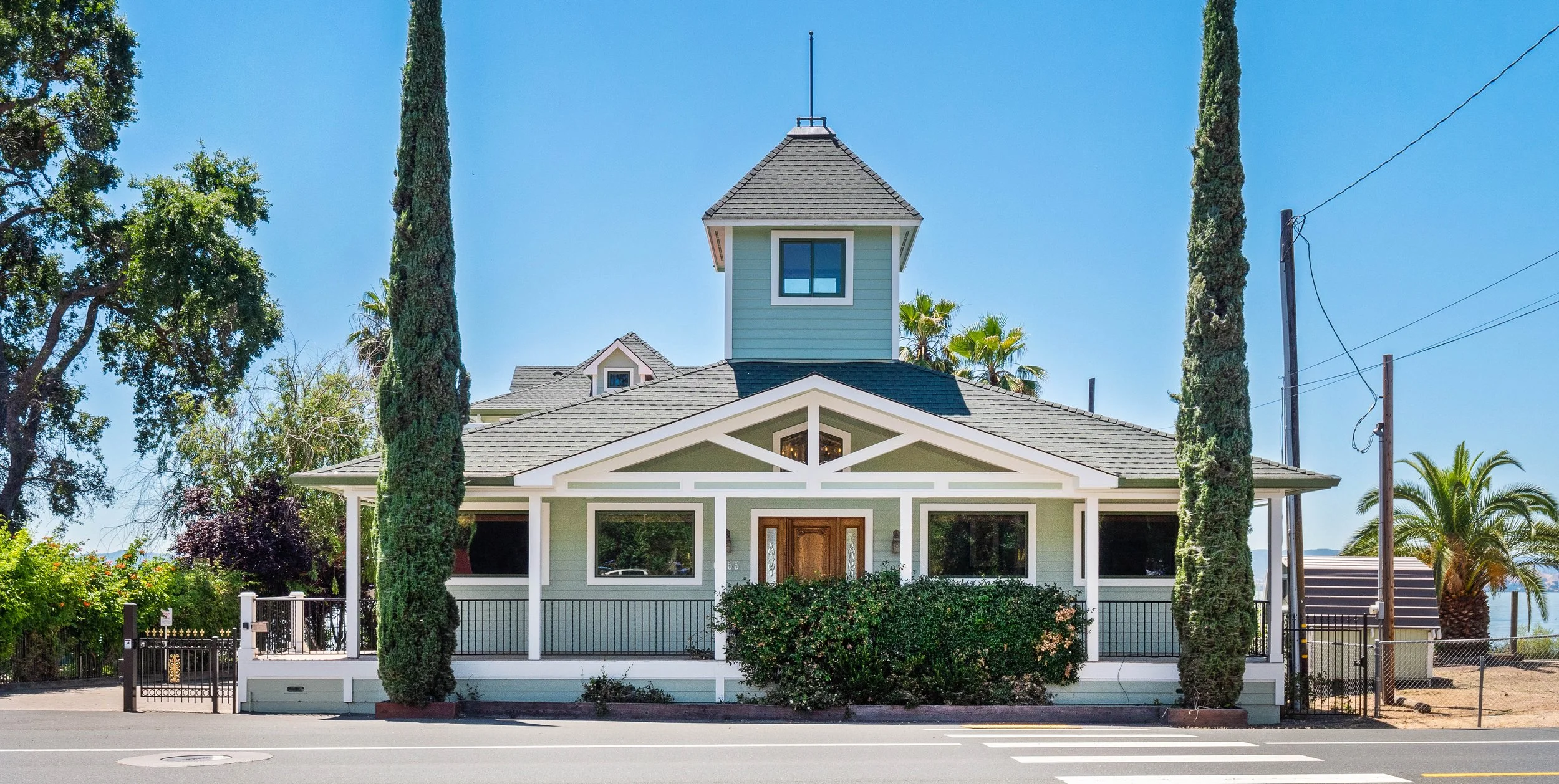 A light blue house with white trim and a small front porch, surrounded by trees, bushes, and a fence. There are utility poles and wires nearby, and the sky is clear and blue.
