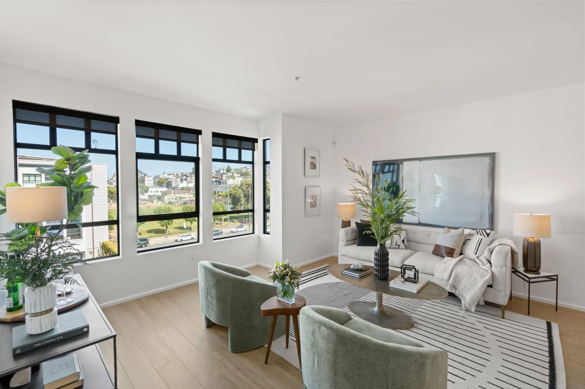 Bright living room with large windows, white walls, a white sofa with decorative pillows, two green armchairs, a wooden coffee table with plants and books, a striped rug, and side lamps.