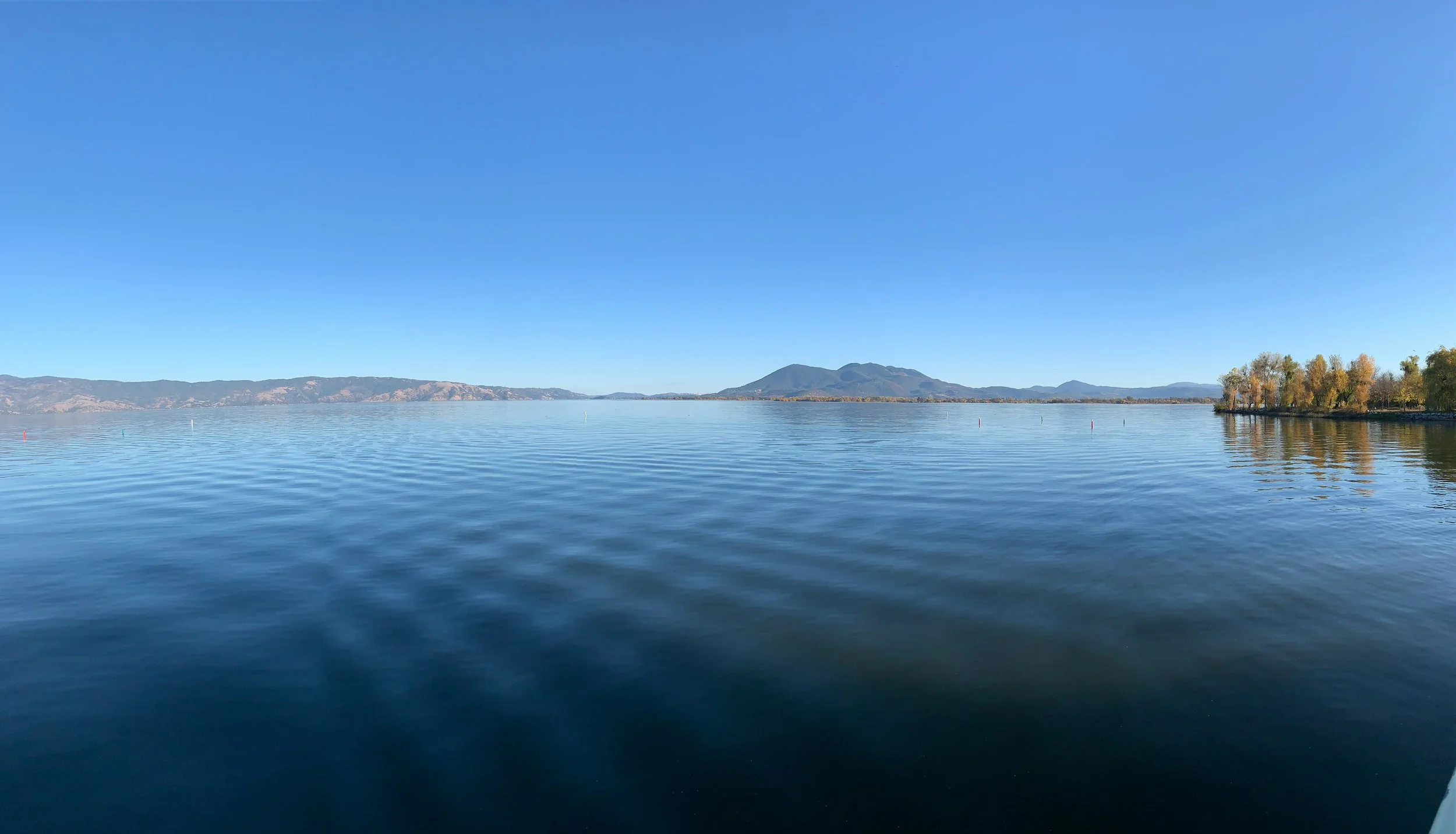 A calm lake with clear blue sky, distant mountains, and trees on the right side with some fall foliage reflected in the water.