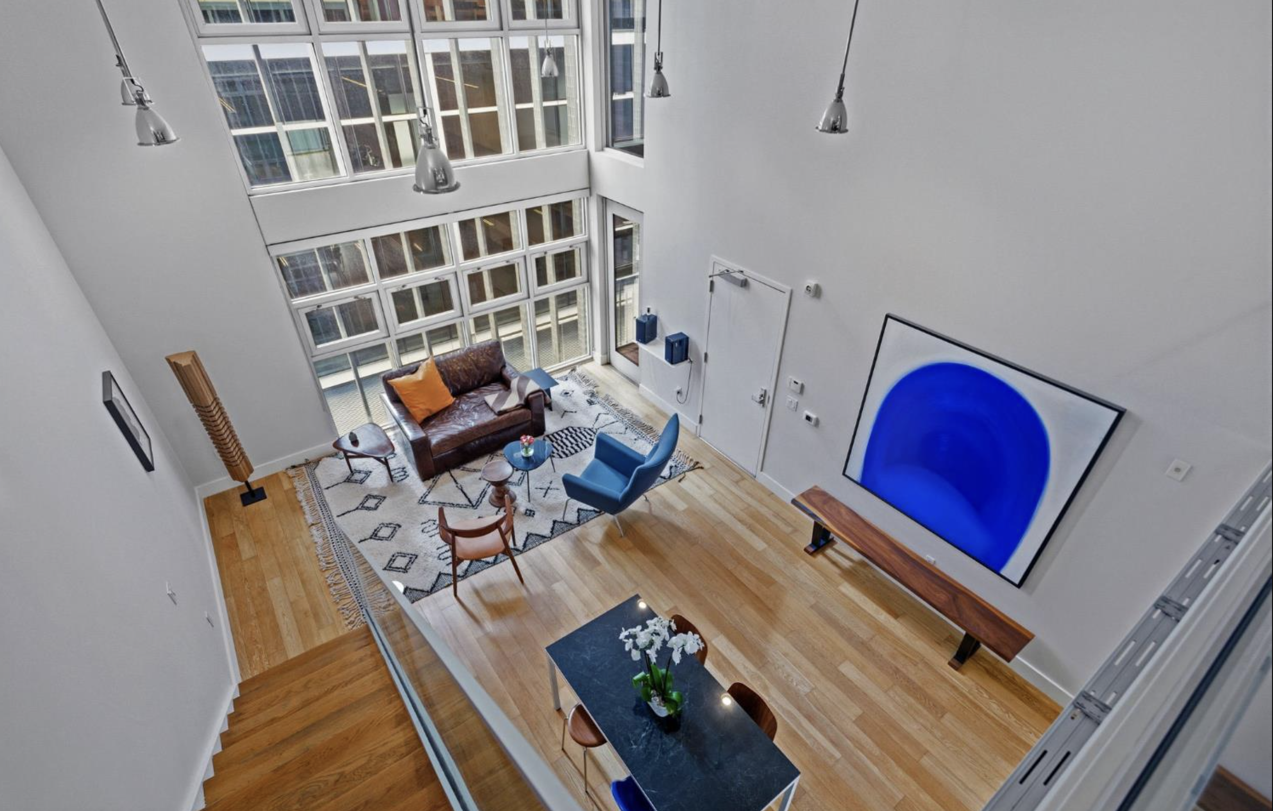 Interior of a modern apartment living room with high ceilings, large windows, a brown leather sofa, a blue armchair, a colorful rug, and a large abstract blue painting on the wall.
