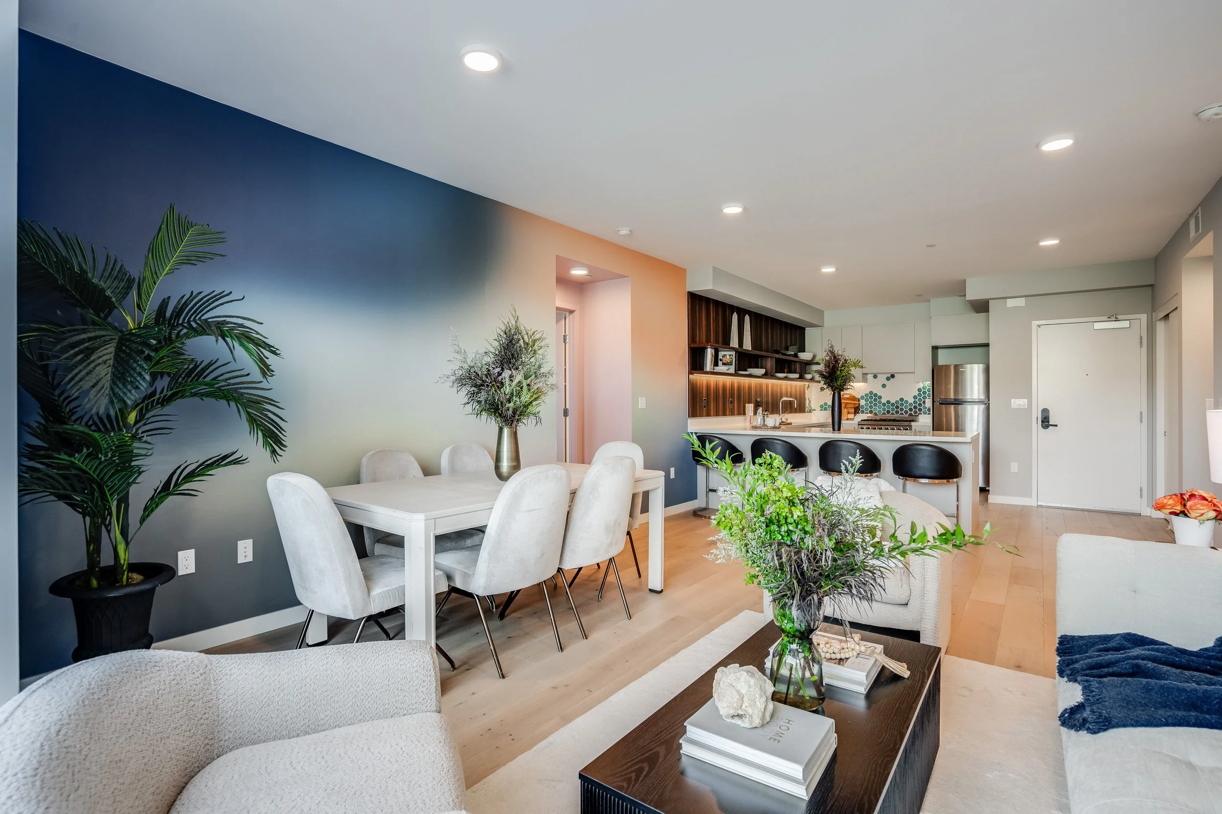 Open-concept living and dining area with light-colored furniture, plants, and a modern kitchen with white cabinets and stainless steel appliances.