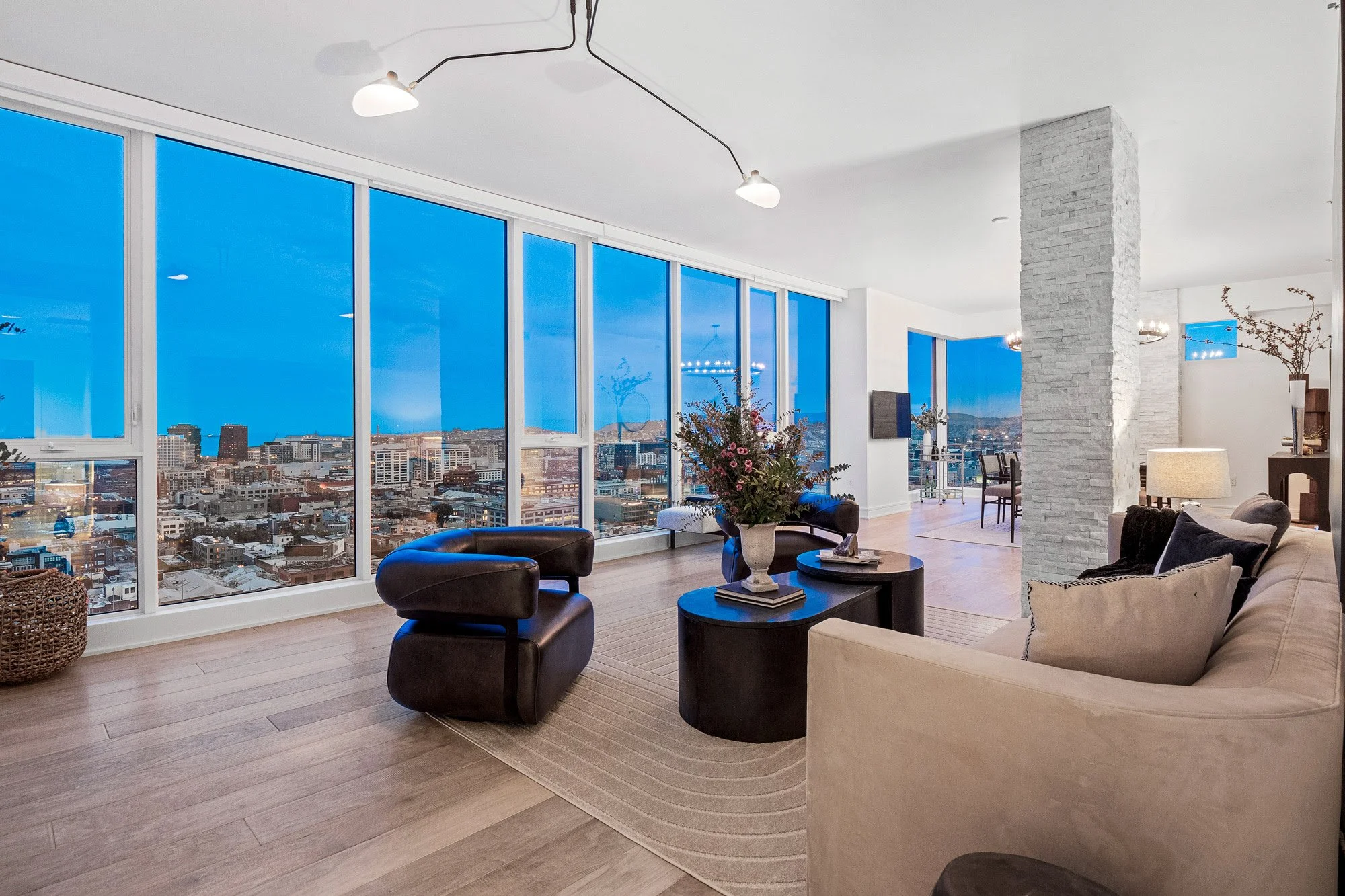 Modern living room with large floor-to-ceiling windows showing a cityscape at dusk, featuring a beige sofa with cushions, black leather armchairs, a coffee table with books and a vase of flowers, a white stone pillar, and a dining area in the background.