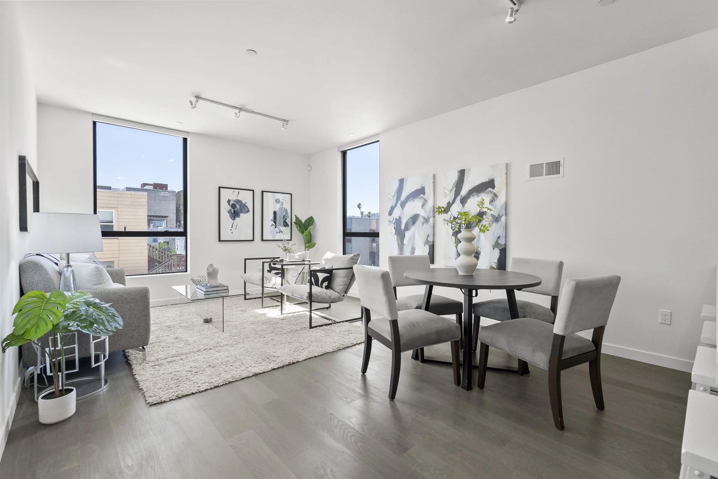 Modern living room with two large windows, light gray sofa, black and white artwork, round dining table with four upholstered chairs, decorative vases, and a beige rug.