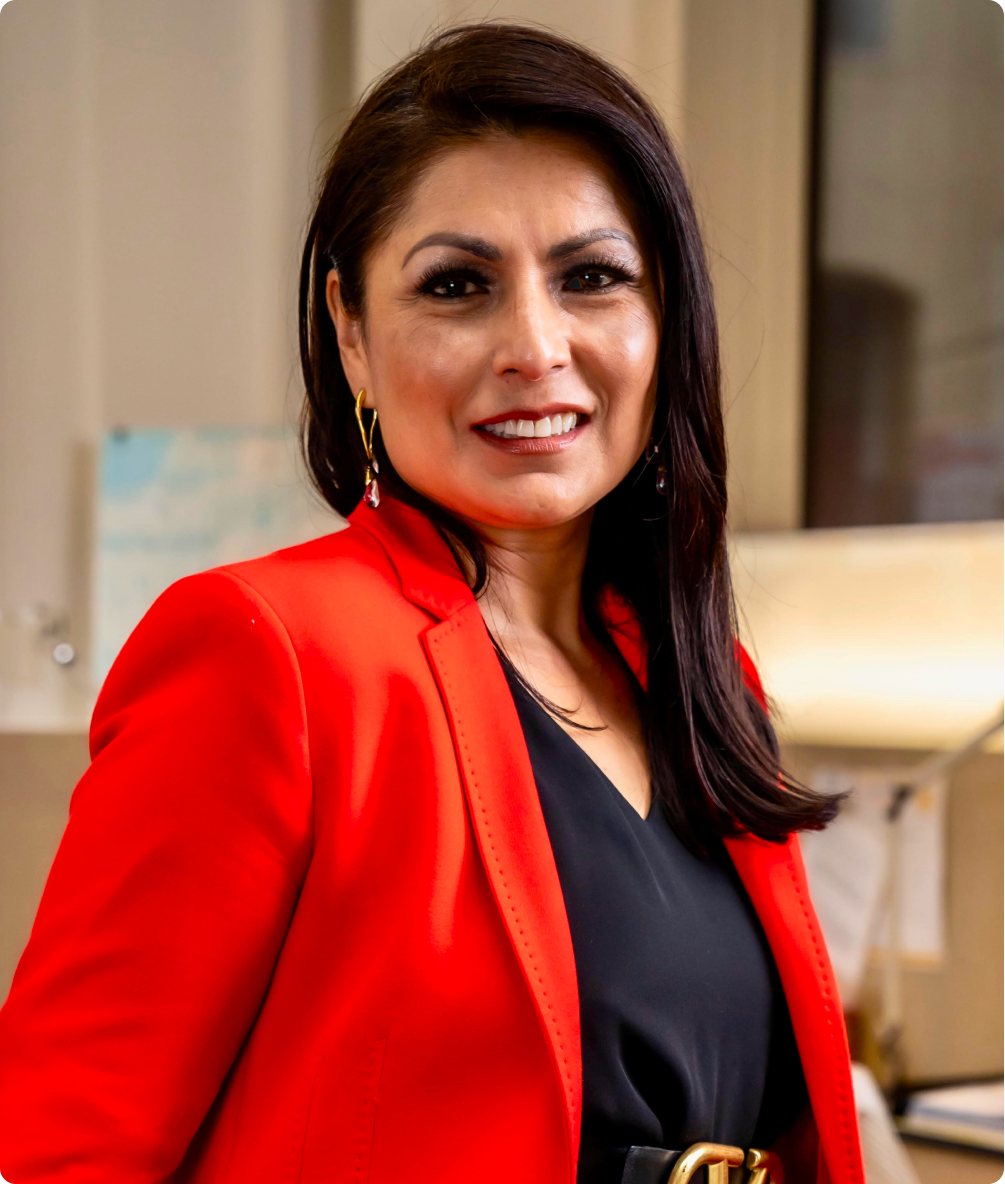A woman with dark brown hair and light makeup, smiling at the camera. She is wearing a red blazer over a black top, with gold earrings. The background appears to be an indoor office or home setting.