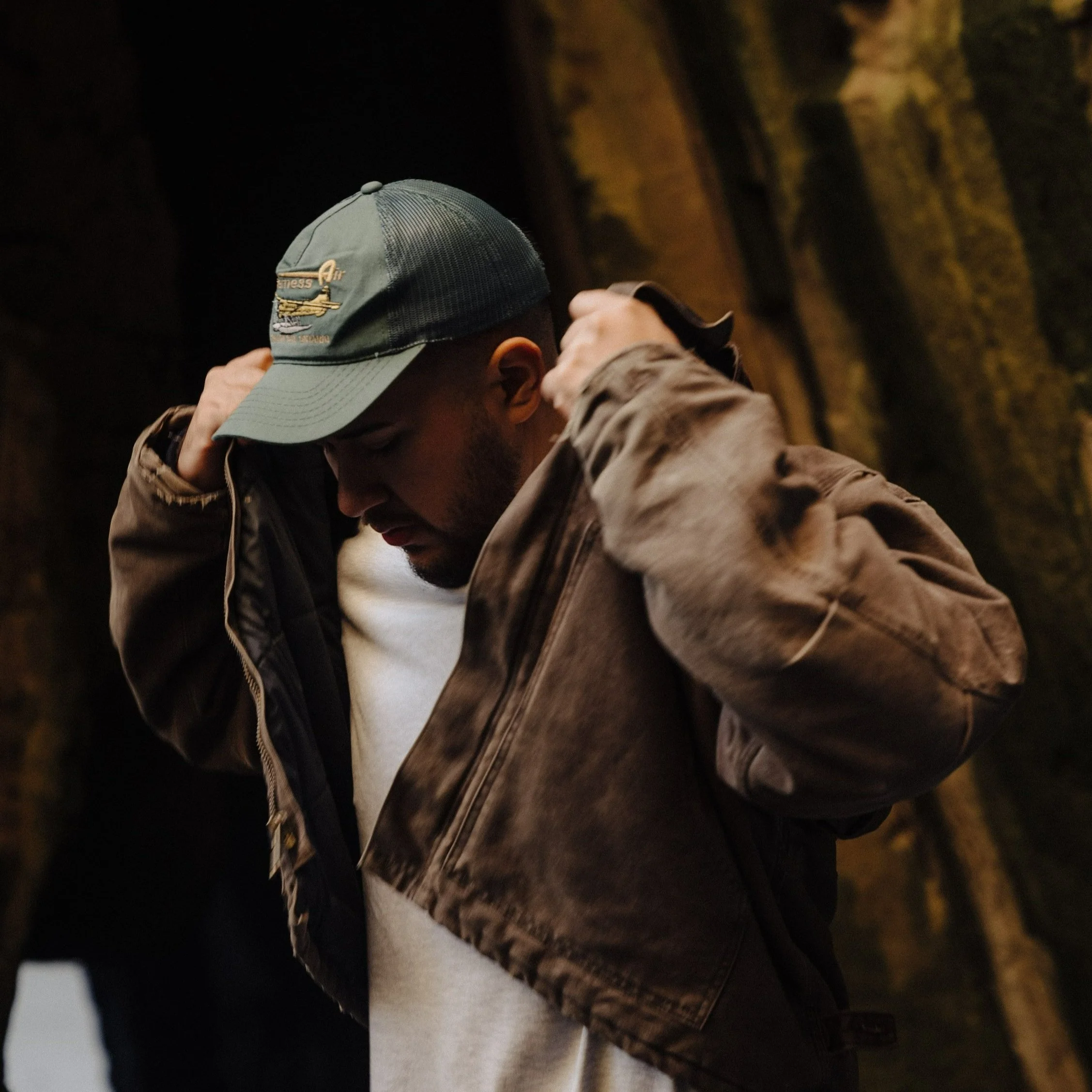 A man wearing a baseball cap, white shirt, and brown jacket, adjusting his jacket with his hands, against a dark background.
