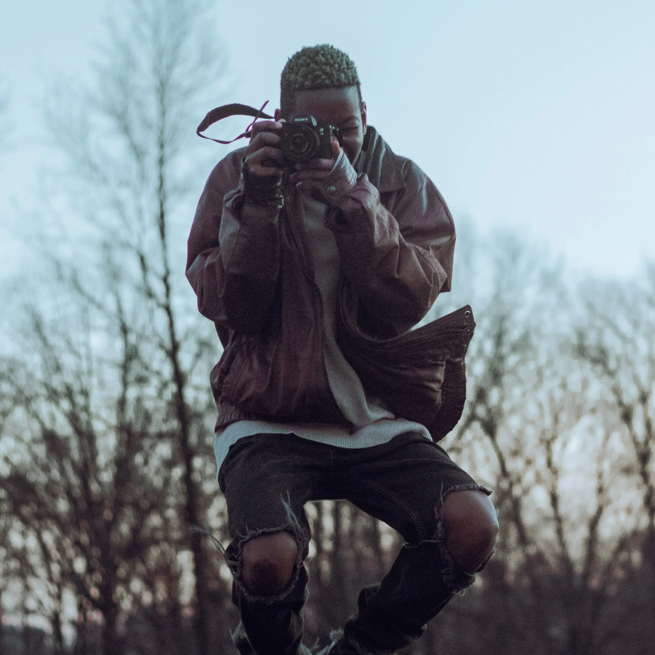 Person in a brown jacket and ripped jeans taking a photo with a camera outdoors during dusk or dawn.