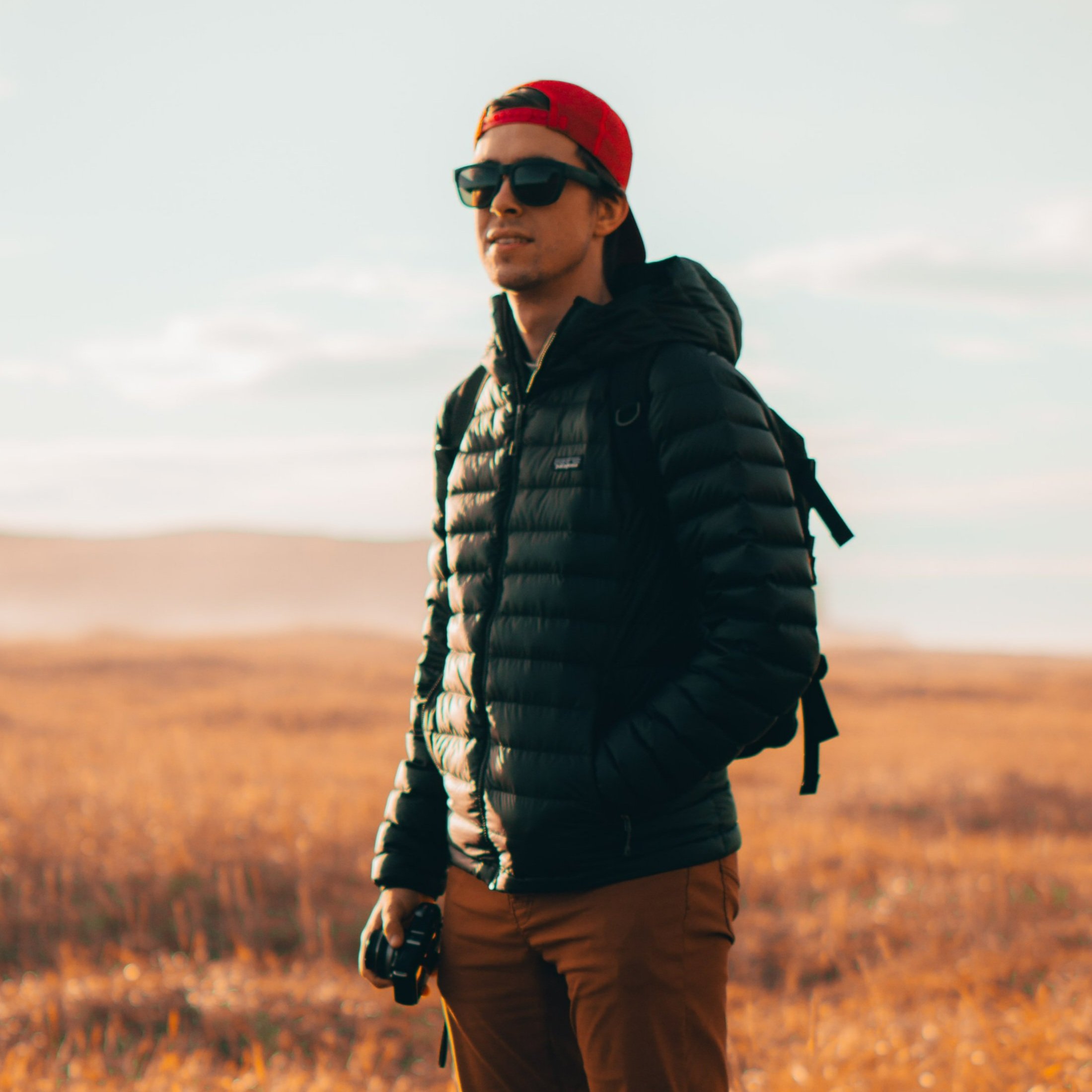 A man standing outdoors in a field with dry grass, wearing sunglasses, a red cap backward, a black puffy jacket, and brown pants, holding a camera in his right hand.