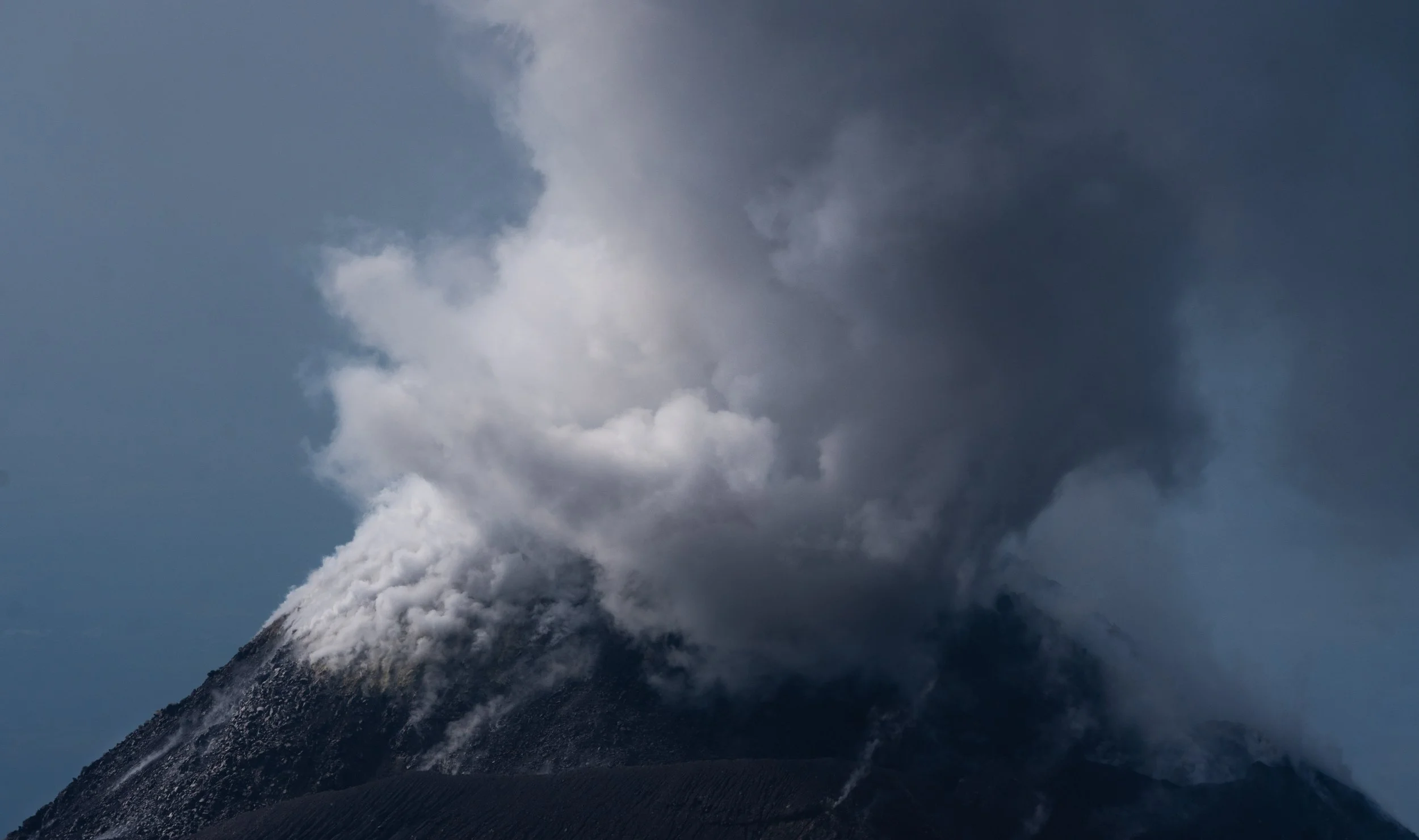 A volcanic eruption with ash and smoke billowing from the summit, dark volcanic rock slopes below.