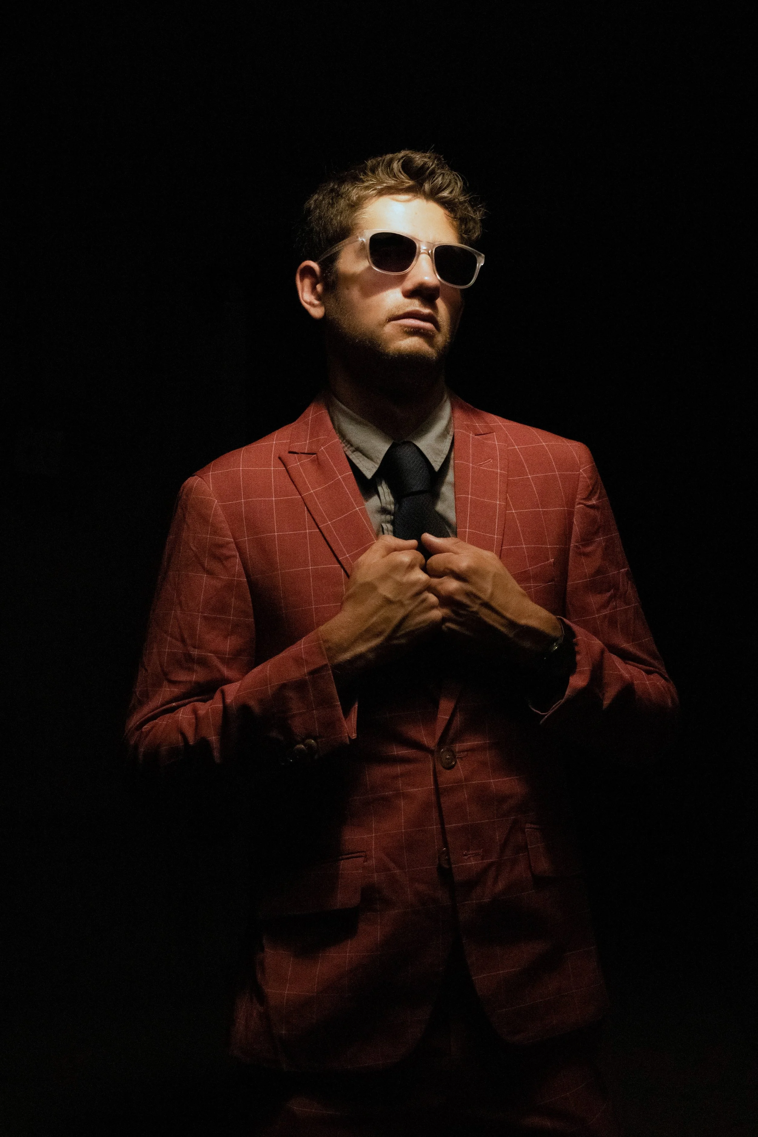 A man in a red checkered suit, sunglasses, and a black tie, posing with his hands adjusting his blazer, against a dark background.
