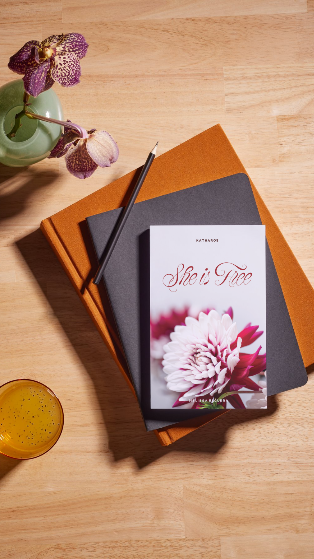 A wooden table with a green vase holding purple orchids, three stacked books with orange, gray, and black covers, and a paperback book titled "She is Free" featuring a flower on the cover. A yellow drink is also partially visible in the bottom left corner.
