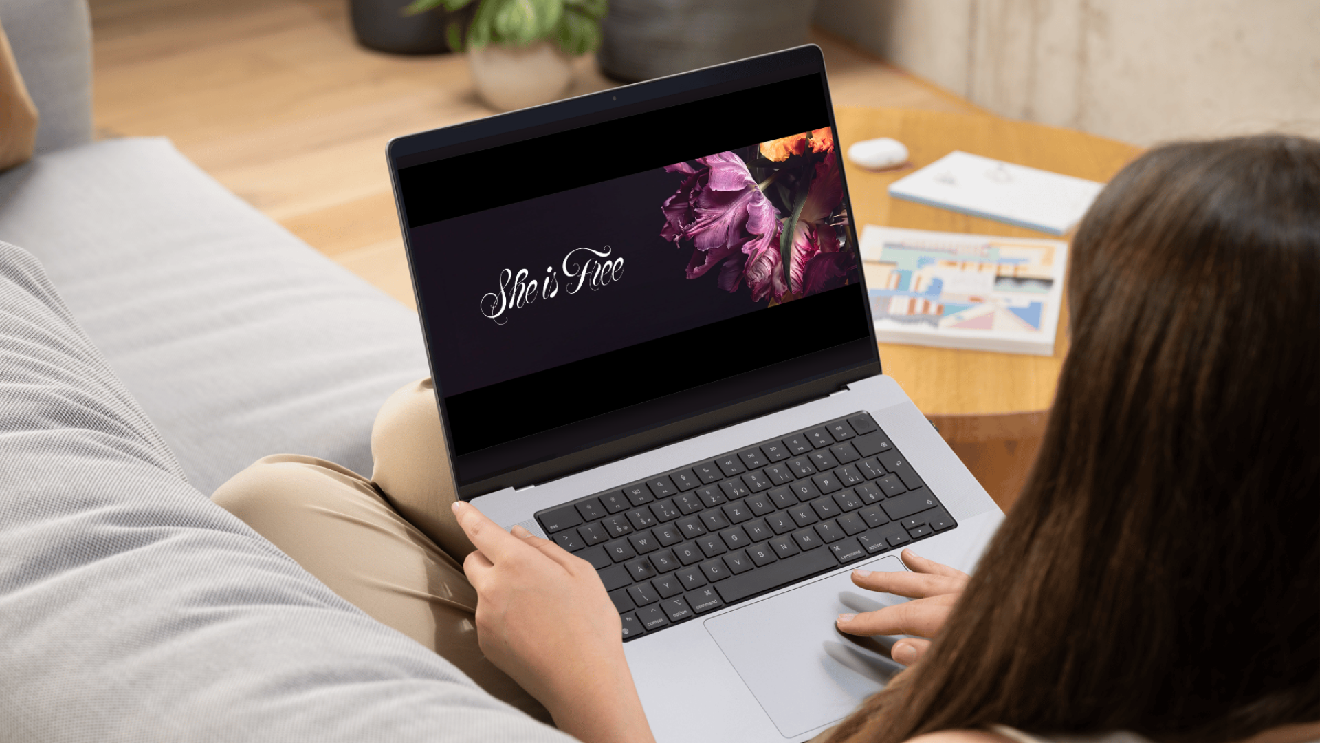 A woman sitting on a couch using a silver laptop with a black keyboard. The laptop screen displays the phrase "She is Free" with purple flowers in the background. A wooden coffee table is in front with a magazine, a white notebook, and wireless earbuds. Potted plants are on the table.