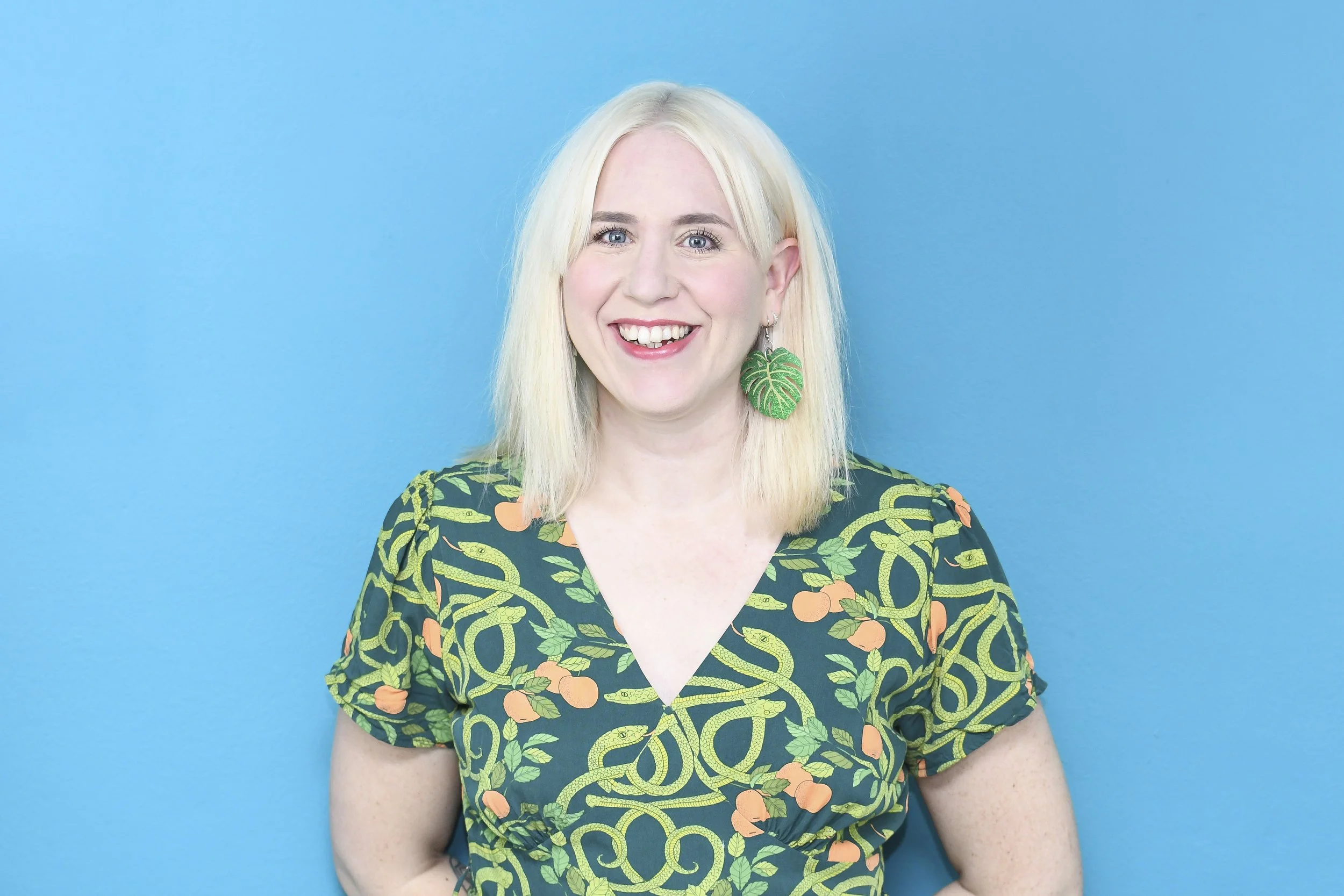 Image of a blonde woman wearing a brightly coloured dress and leaf design earrings. She is standing in front of a light blue background.