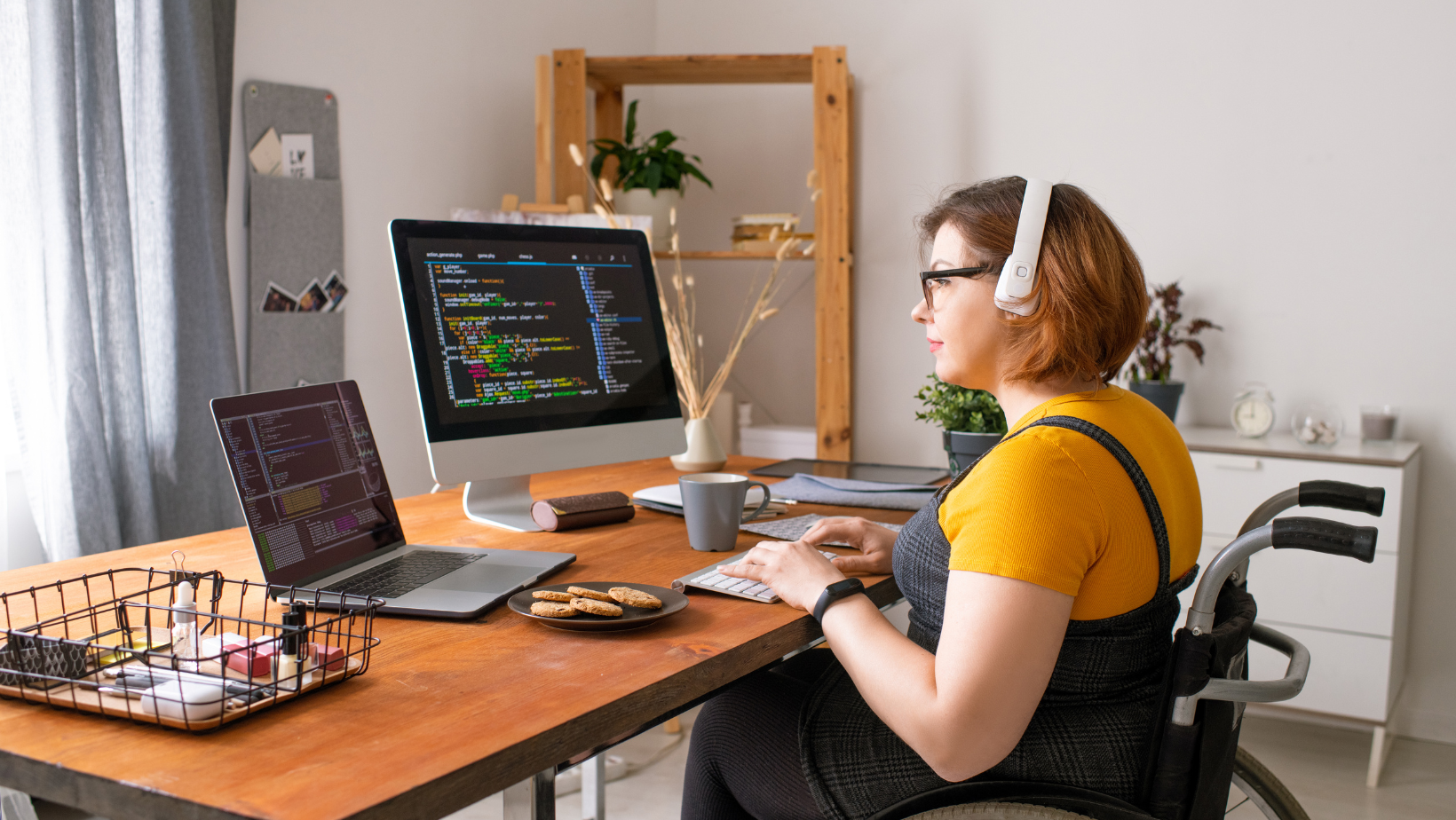 A white woman with short brown hair sat at a desk with a laptop and a computer screen. She is wearing black dungarees and a yellow top as well as noise cancelling headphones. She is sat in a manual wheelchair.