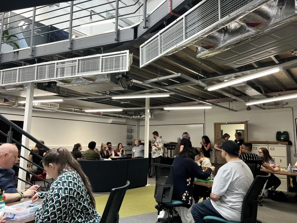 A picture taken during a LEGO serious play workshop. People sat at tables are either focusing on their LEGO or looking at Dan, the workshop facilitator. They're all in an atrium space with a balcony over them.