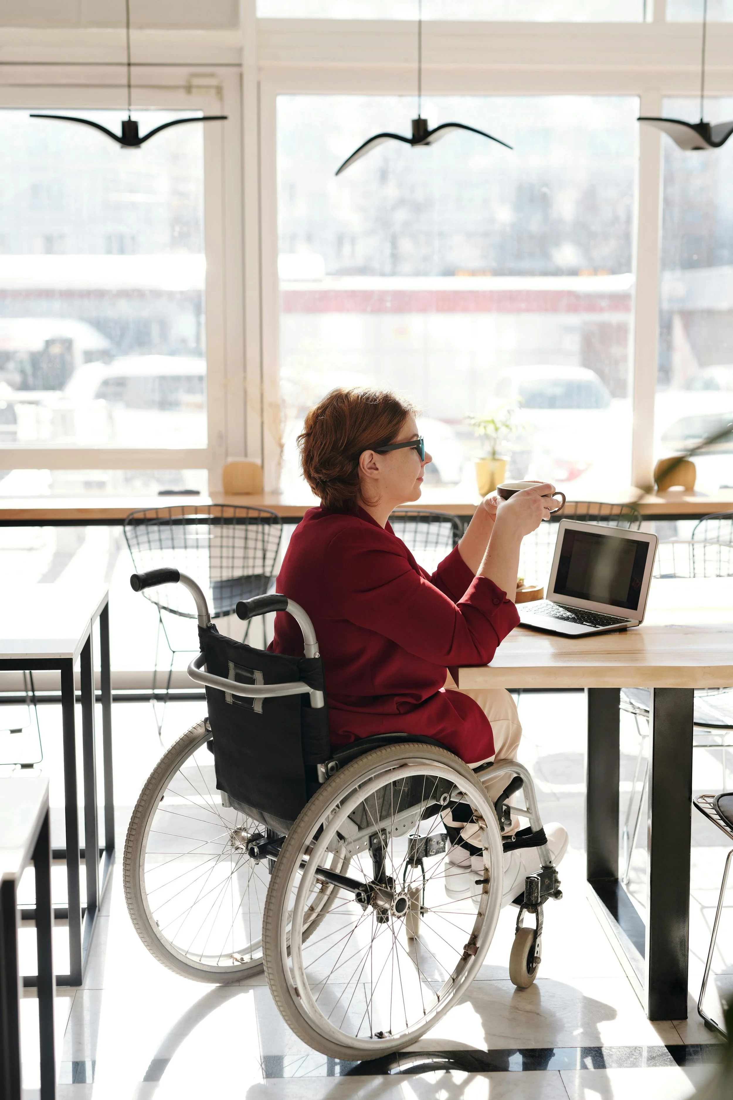 A woman in a wheelchair sitting at a desk and drinking a coffee. DShe has short dark hair and is wearing a dark red jumper.