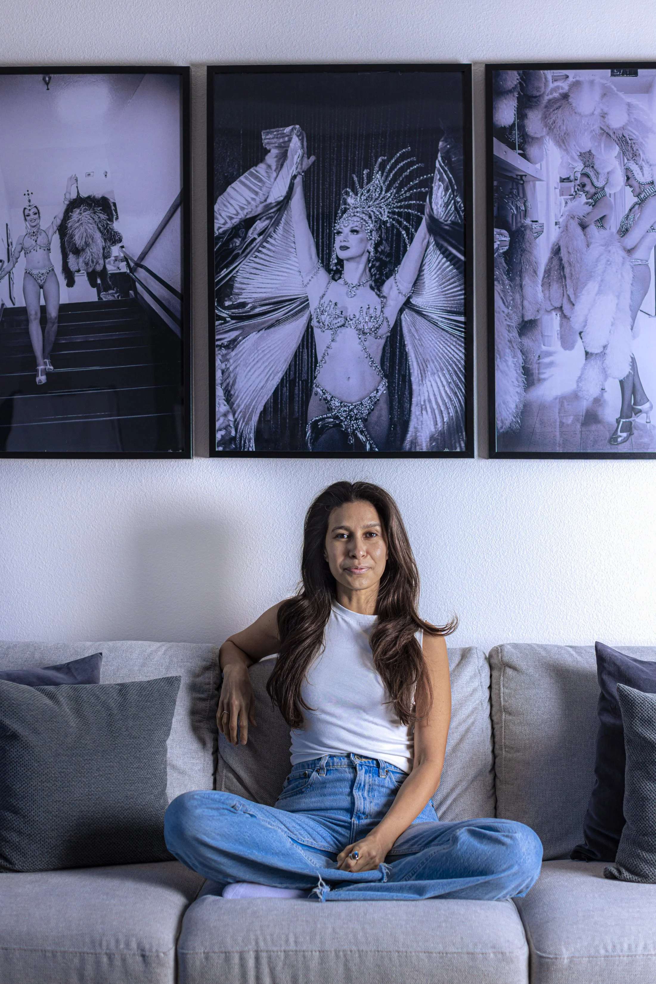 The founder of The Tidy Armadillo, Jessi, is a woman with long dark hair, wearing a white sleeveless top and blue jeans, sitting cross-legged on a gray couch in front of three large black and white photographs of showgirls in costumes.