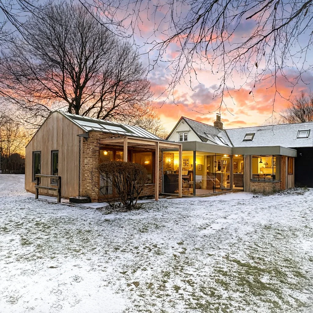 A house with a modern extension featuring large windows and a glass roof, illuminated from within, set in a snow-covered yard with leafless trees at sunset.