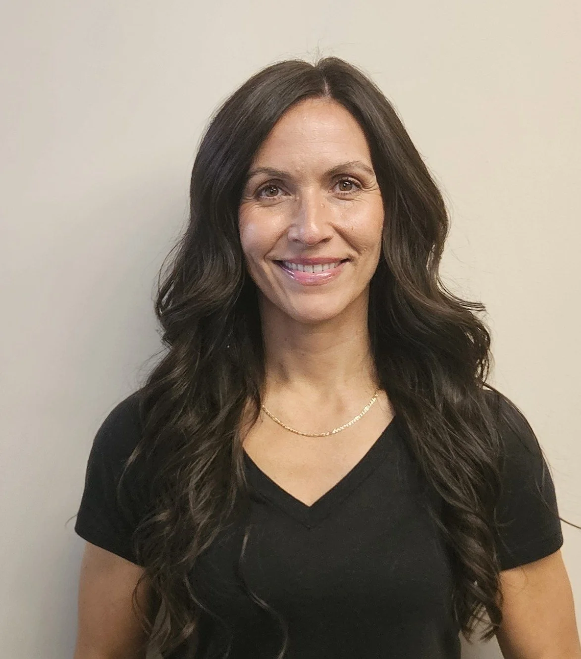 A woman, Chandra, with long dark wavy hair smiling, wearing a black V-neck shirt and a gold chain necklace, standing against an off-white wall.