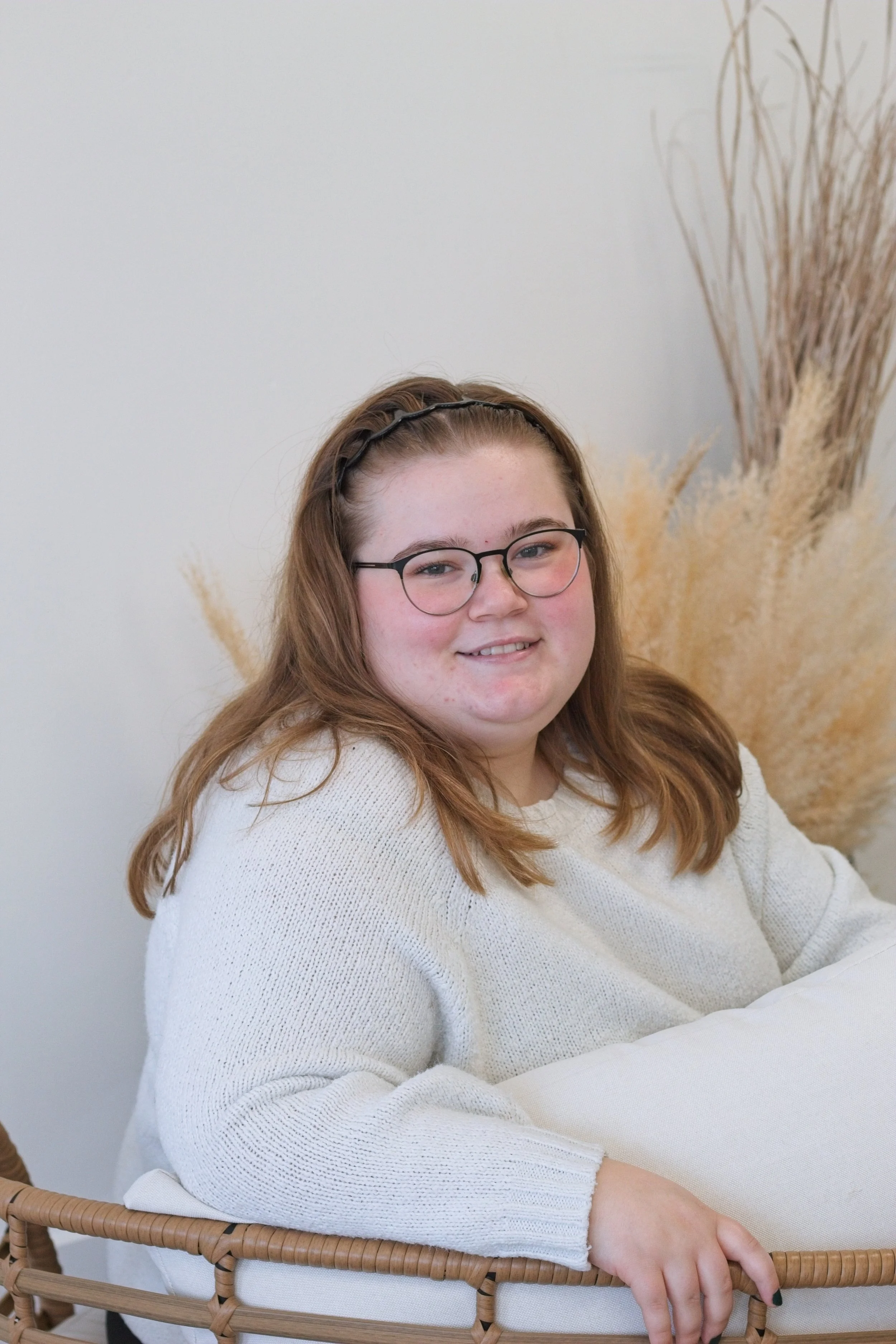 A young woman with glasses and auburn hair sitting on a wicker chair in front of a light-colored background with tall, beige dried plants.