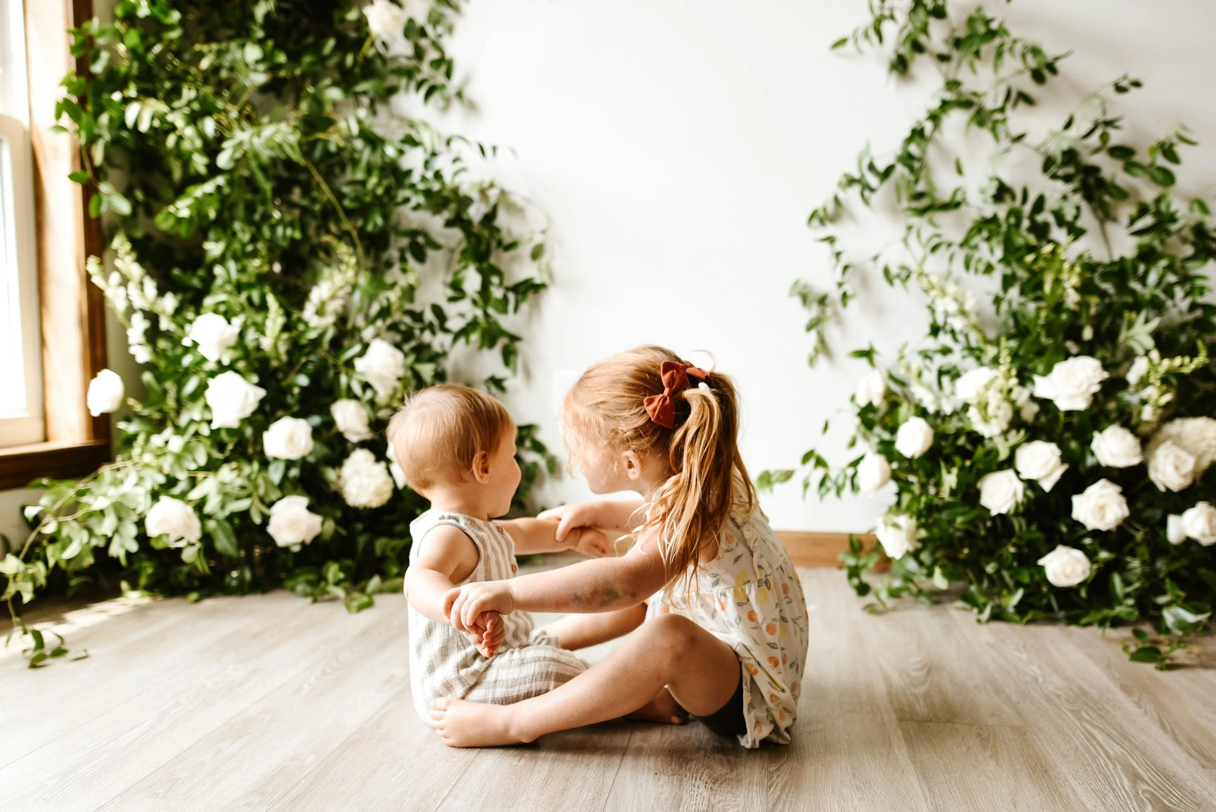 Two young children, a girl and a boy, sitting on a wooden floor surrounded by large white flowers and green foliage, engaging playfully with each other.