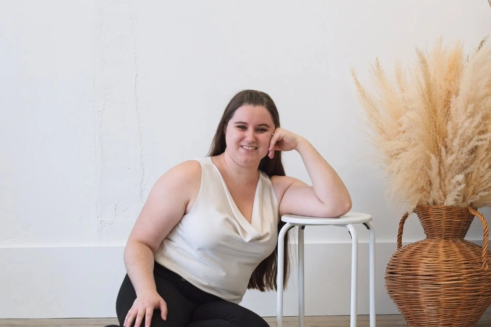A woman with long brown hair sitting on the floor next to a small white table and a large wicker vase with dried pampas grass, smiling at the camera.