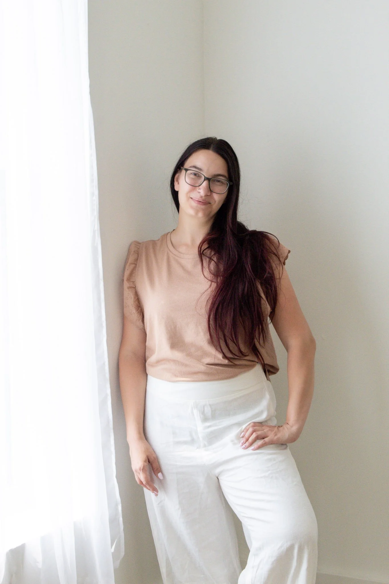 A woman with long dark hair and glasses, smiling, standing barefoot on a wooden floor with a plain white wall behind her, wearing a green dress.