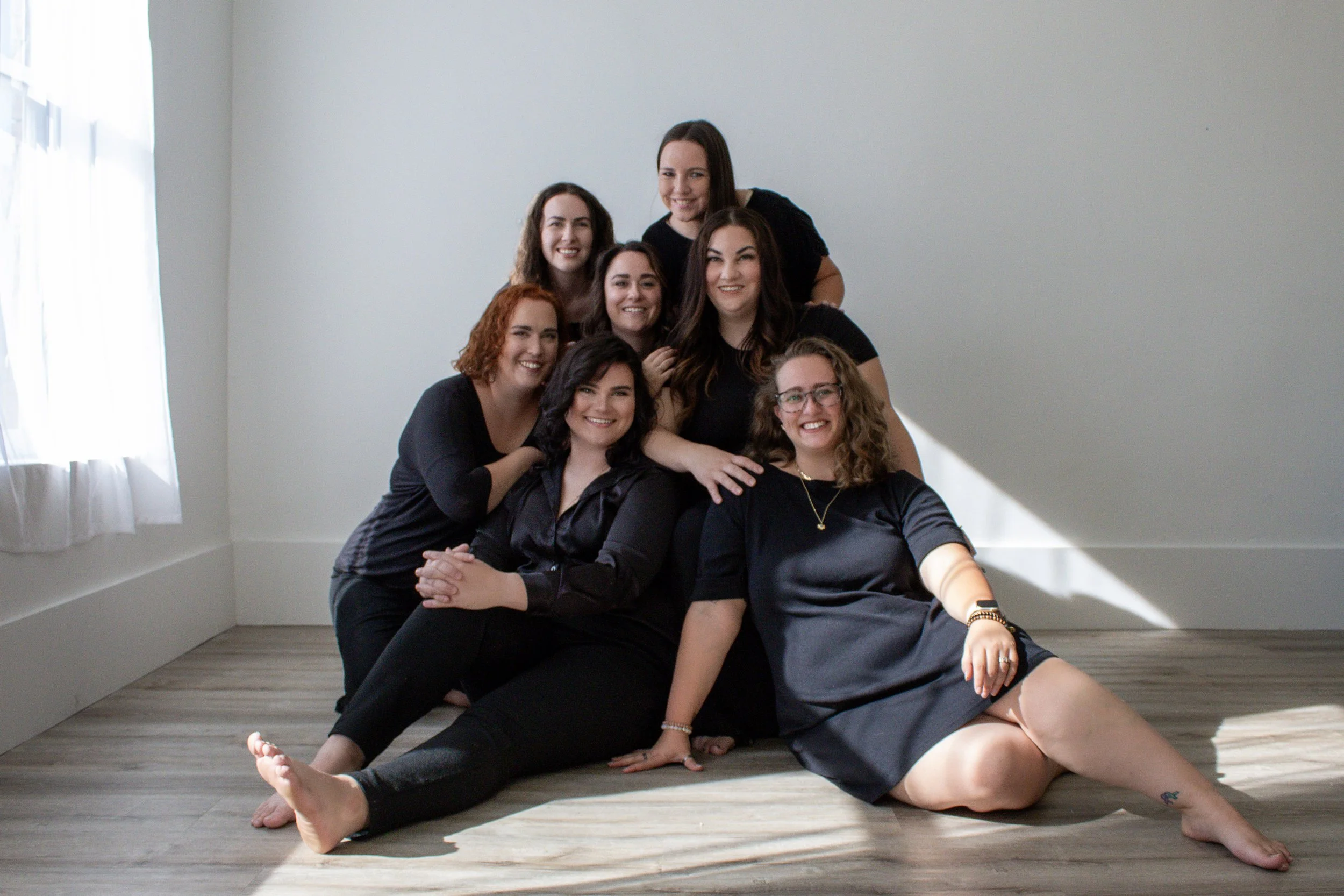 Eight women in black clothing sitting and standing on a light wood floor in a bright room with white walls and a white curtain, smiling for a group photo.