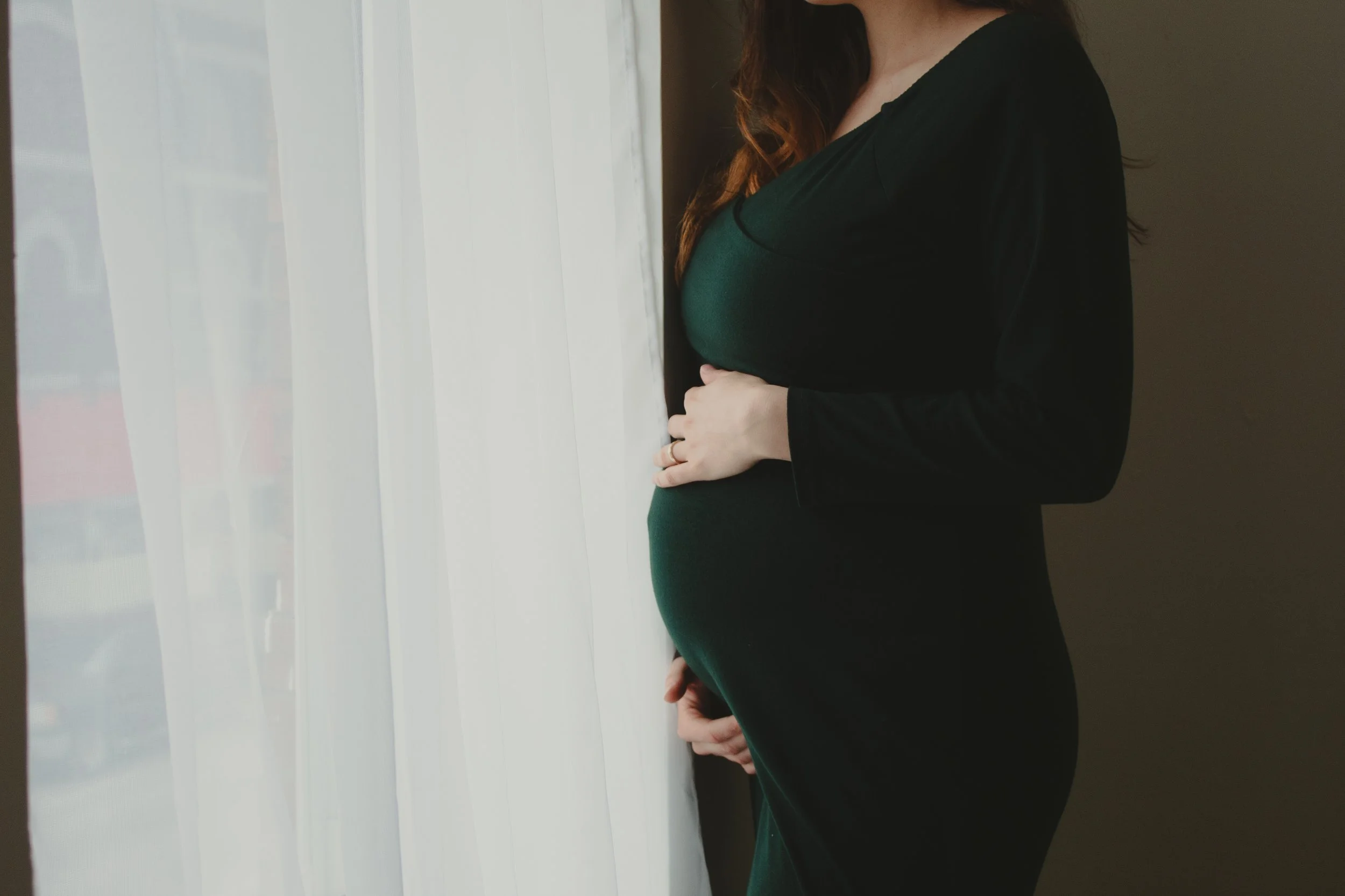 A pregnant woman in a black long-sleeve dress standing by a window with sheer white curtains, gently holding her baby bump.