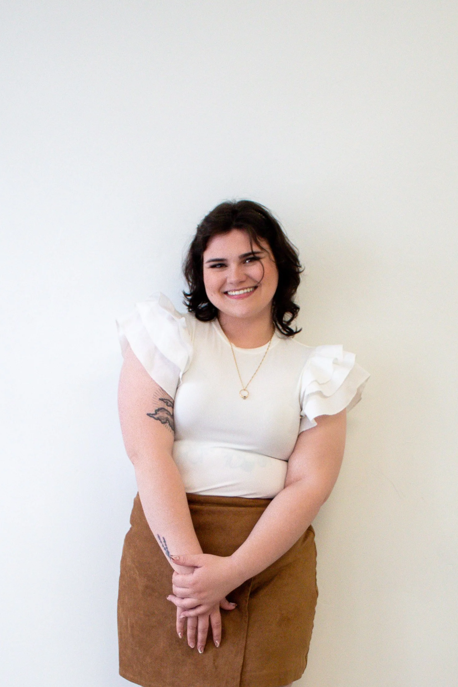 Young woman with dark hair smiling, wearing a white ruffled-sleeve top, brown skirt, and a necklace, standing against a plain white wall.