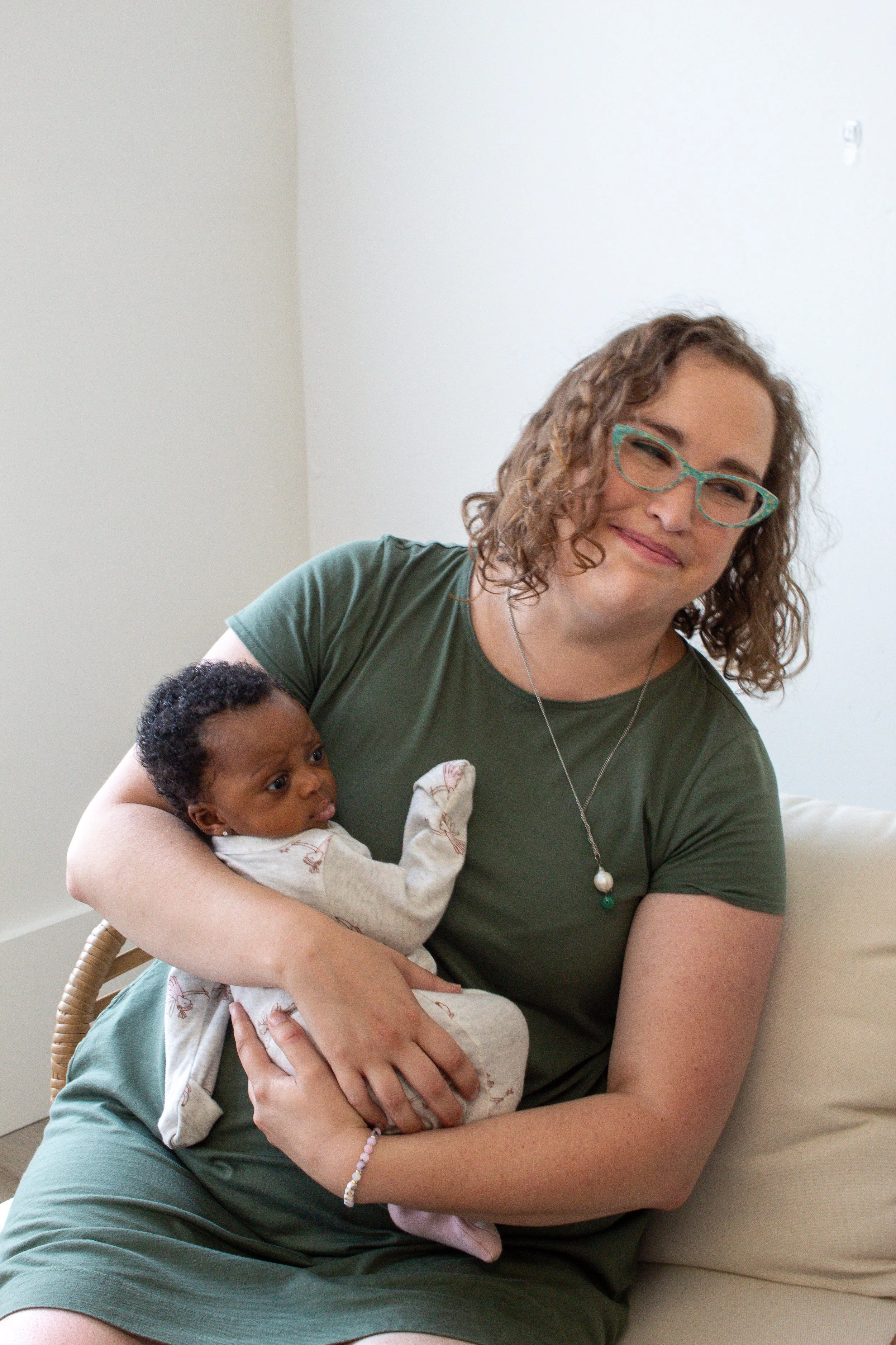 A woman with curly hair and glasses holding a baby with short curly hair and dark skin. They are seated indoors against a plain wall with natural light.