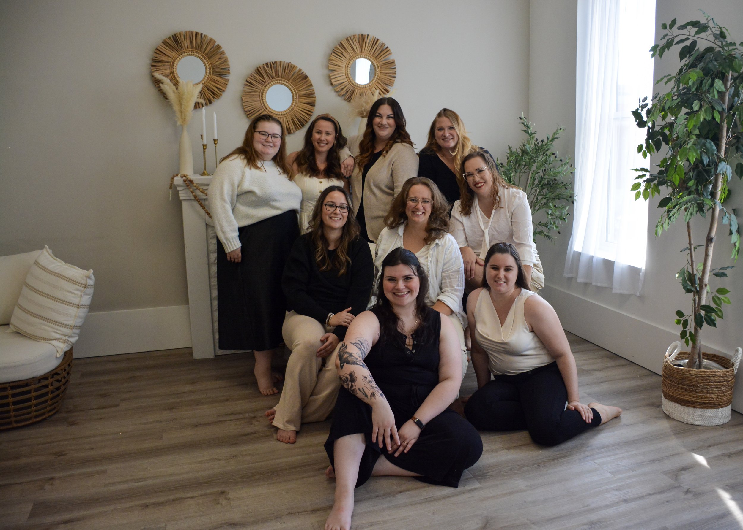 Group of women gathered in a cozy, well-lit room with decorated wall mirrors, plants, and neutral decor, smiling at the camera.