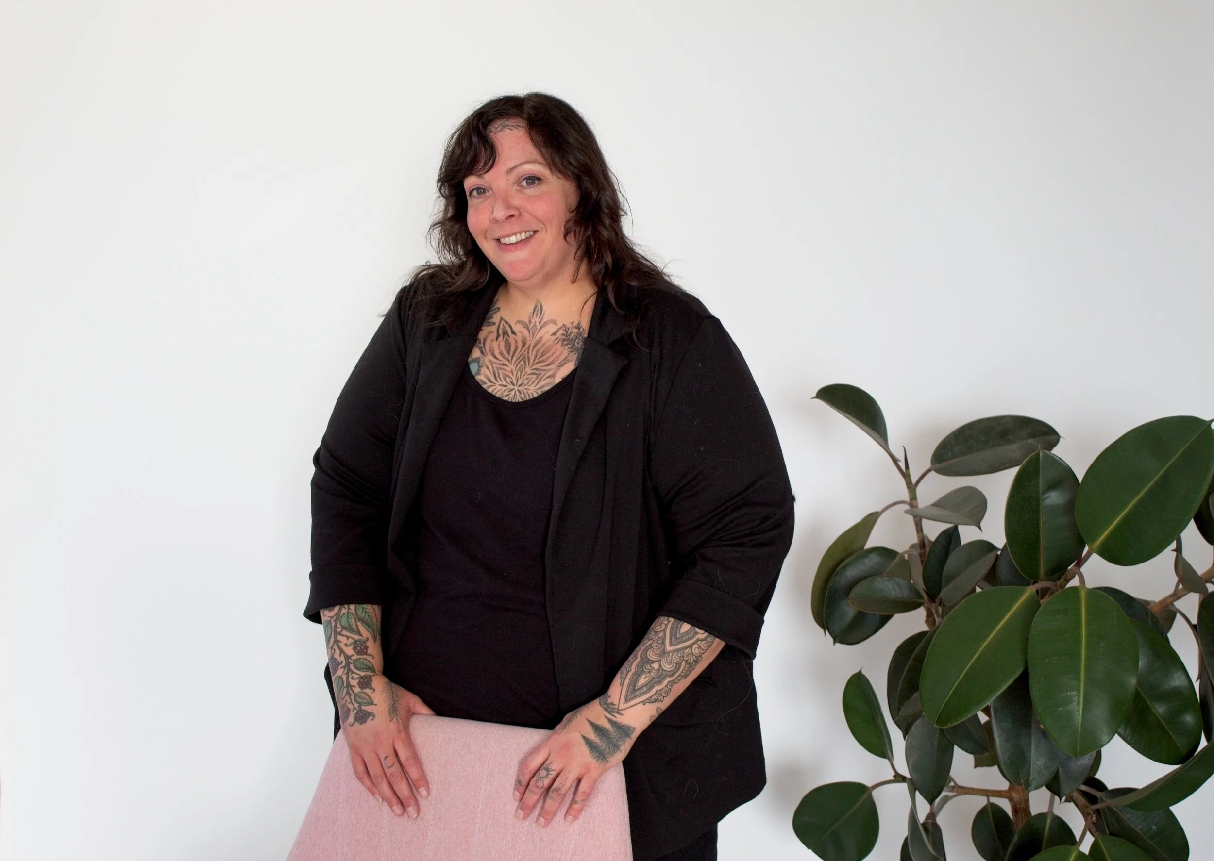 A woman with dark, wavy hair and tattoos on her arms and chest, wearing a black jacket and shirt, standing and leaning slightly on a pink chair, smiling at the camera, with a large green leafy plant beside her against a plain white background.
