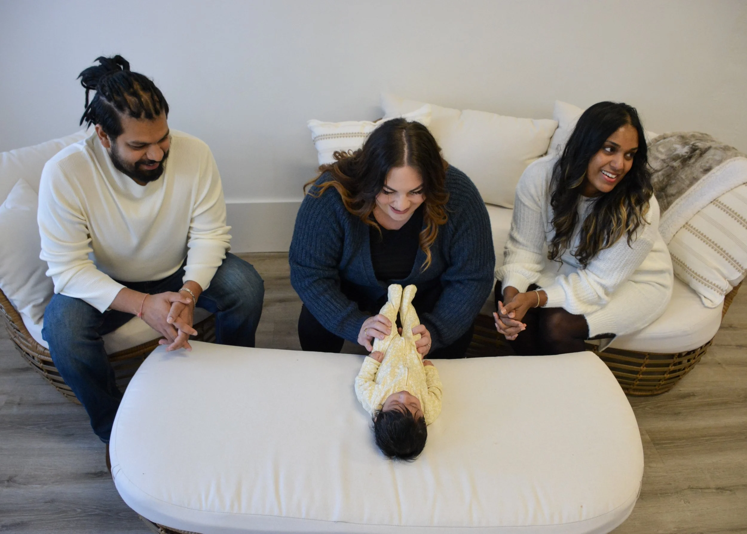 A woman holding a baby on a changing table while two people, a man and a woman, look on and smile in a cozy living room.