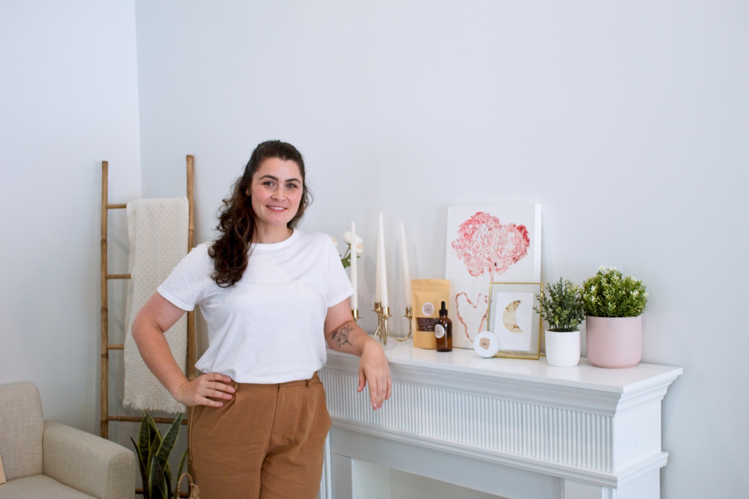 A woman with brown hair and a white t-shirt stands near a white fireplace with decorative items including candles, framed artwork, plants, and a small bottle. She has her hand on her hip and is smiling.