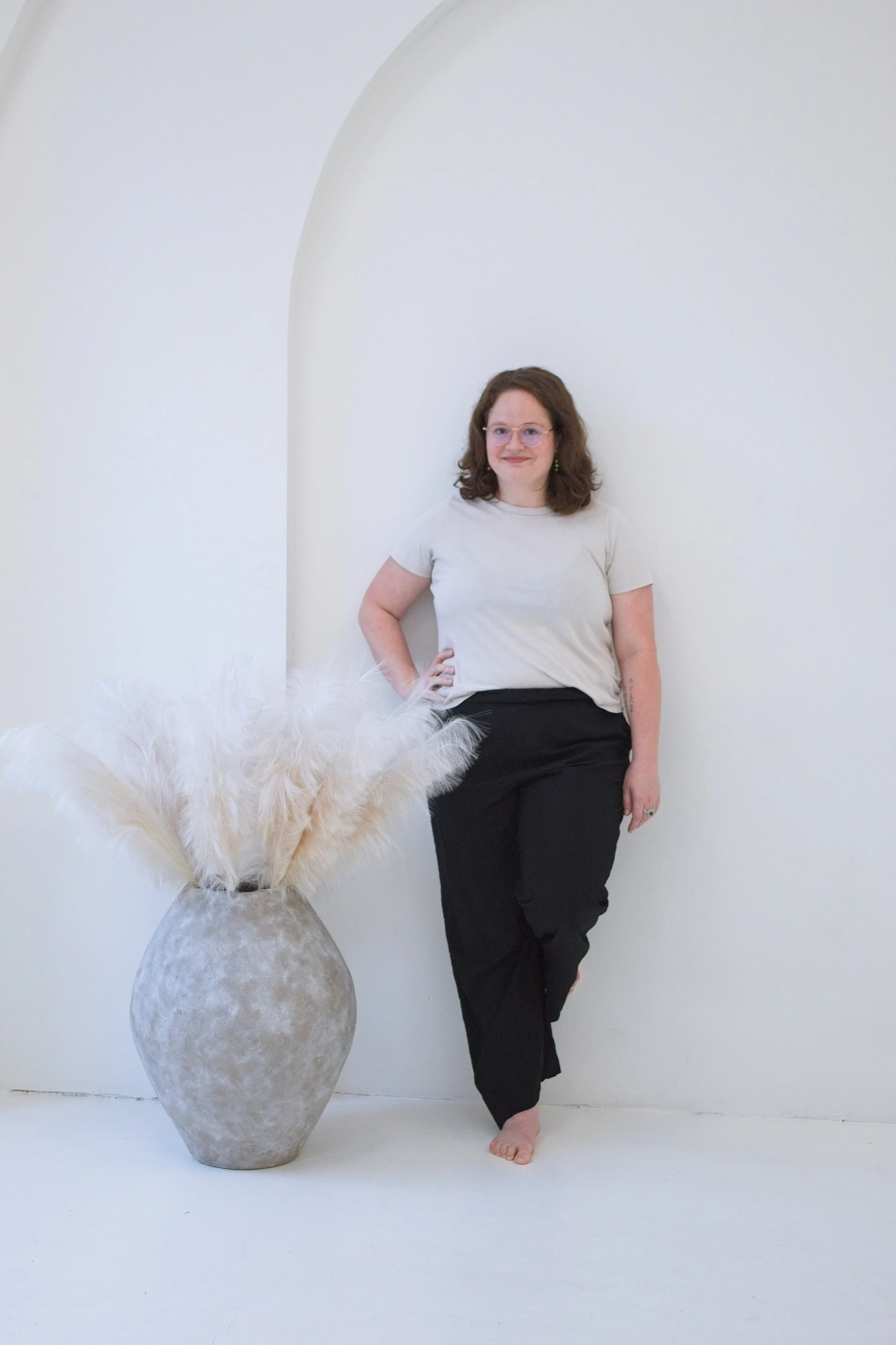 A woman with curly brown hair, glasses, and tattoos stands barefoot next to a large textured vase with white pampas grass, against a white wall with an arch opening.