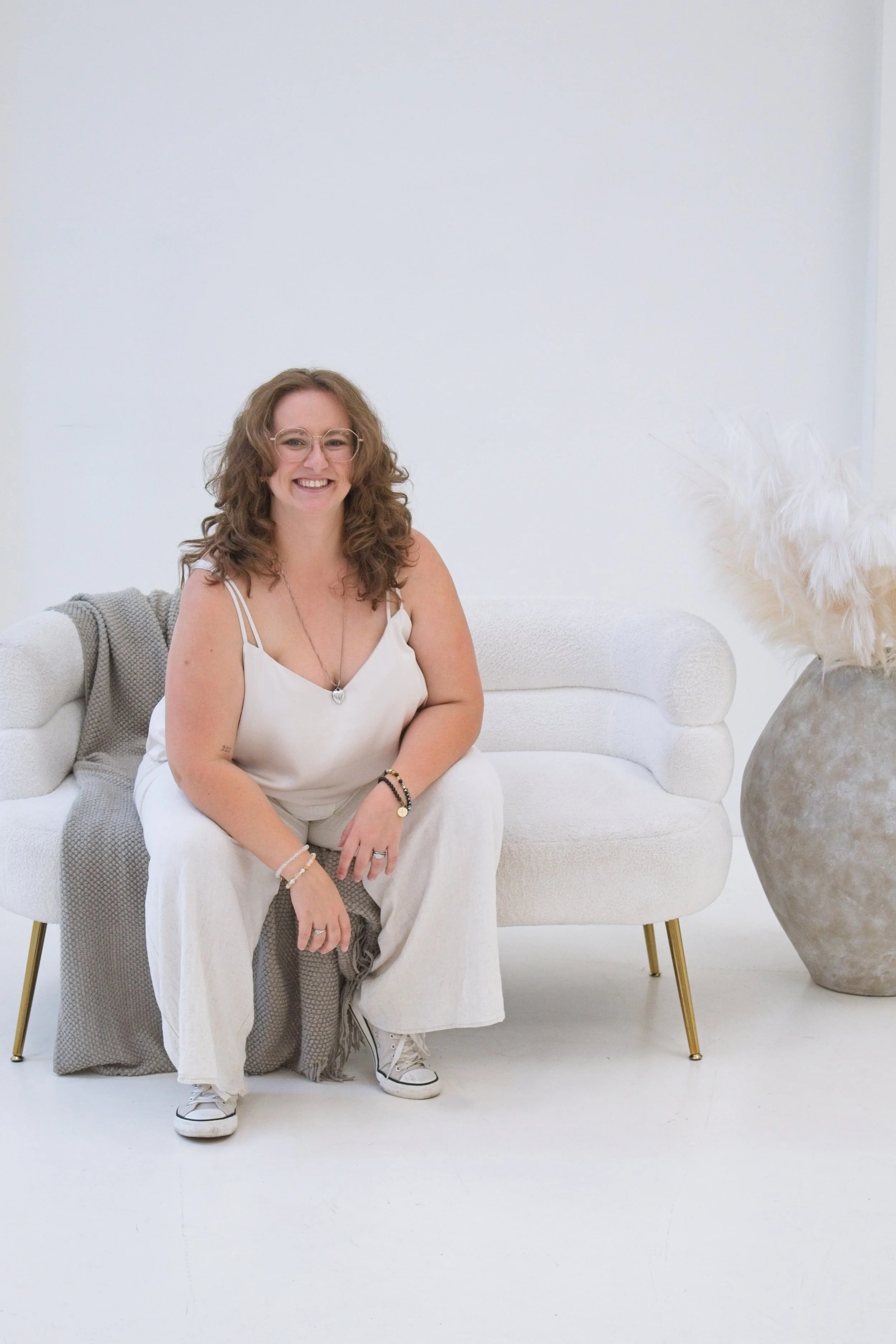 Woman with curly hair and glasses sitting on a white couch in a minimalist room with white walls and a large vase with pampas grass.
