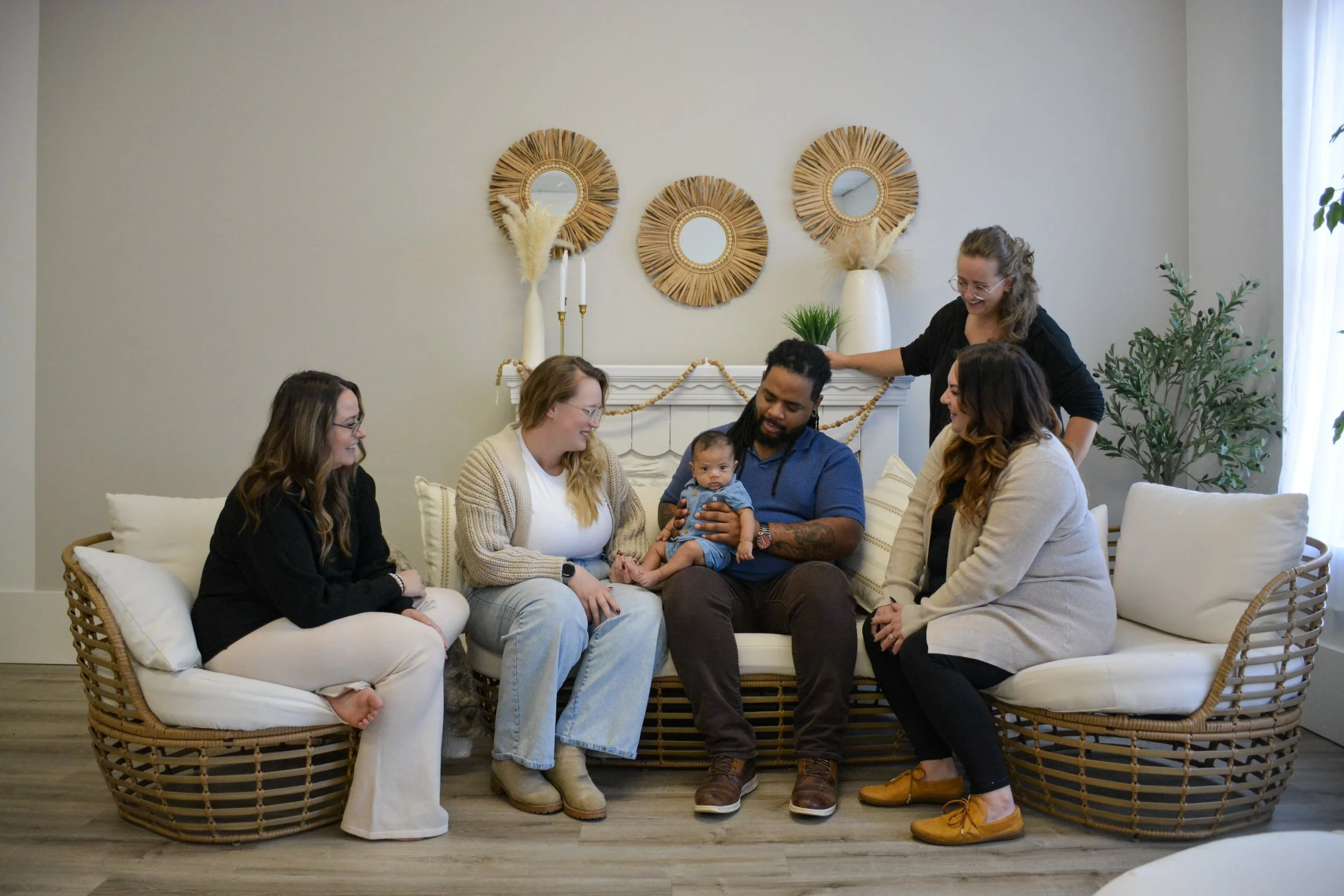 Group of people sitting on a sofa in a living room, celebrating a baby's first birthday, with birthday decorations on the wall.