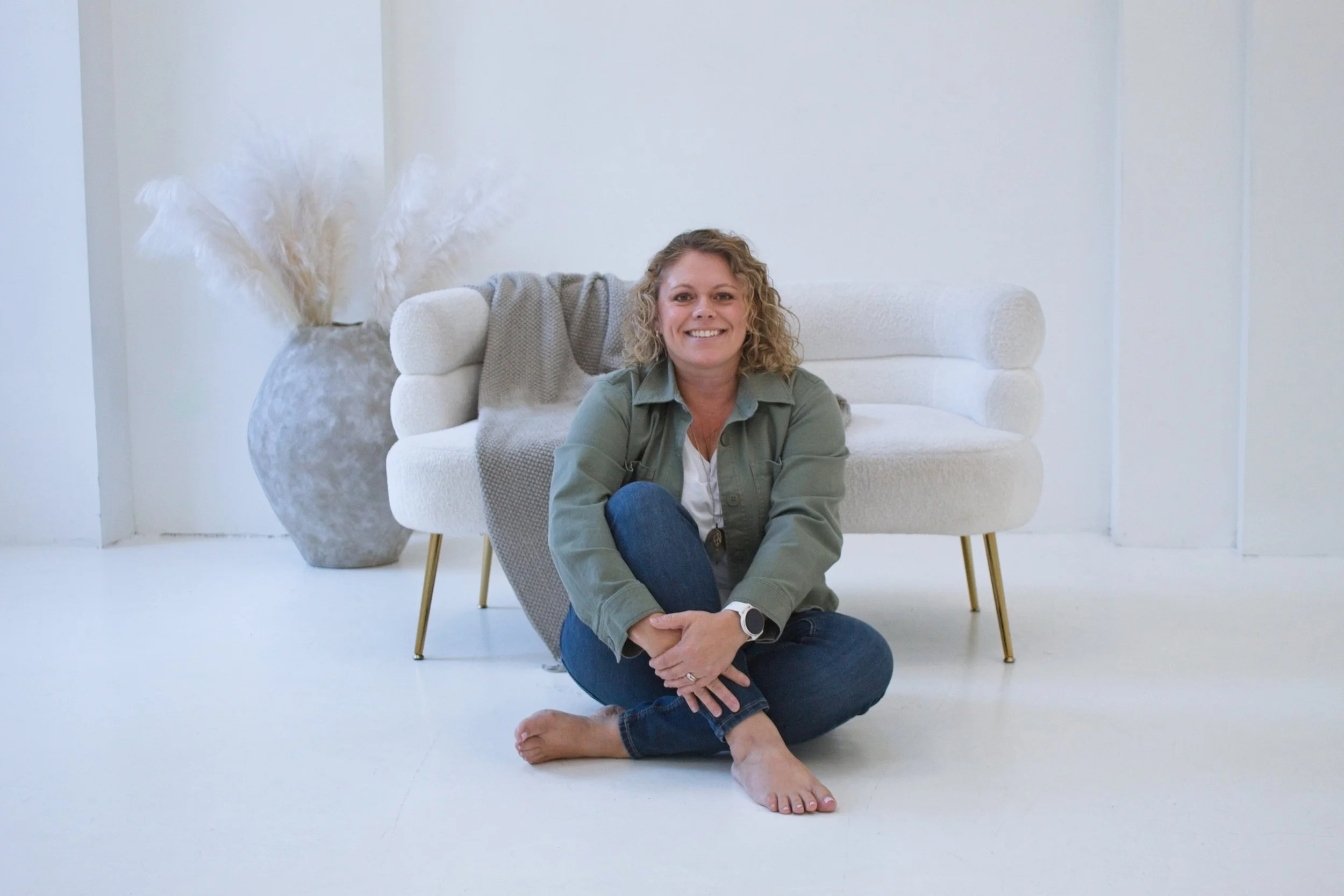 A woman with curly blonde hair wearing a green jacket and blue jeans, sitting on the floor barefoot, smiling at the camera with a white sofa and a large vase with pampas grass behind her.