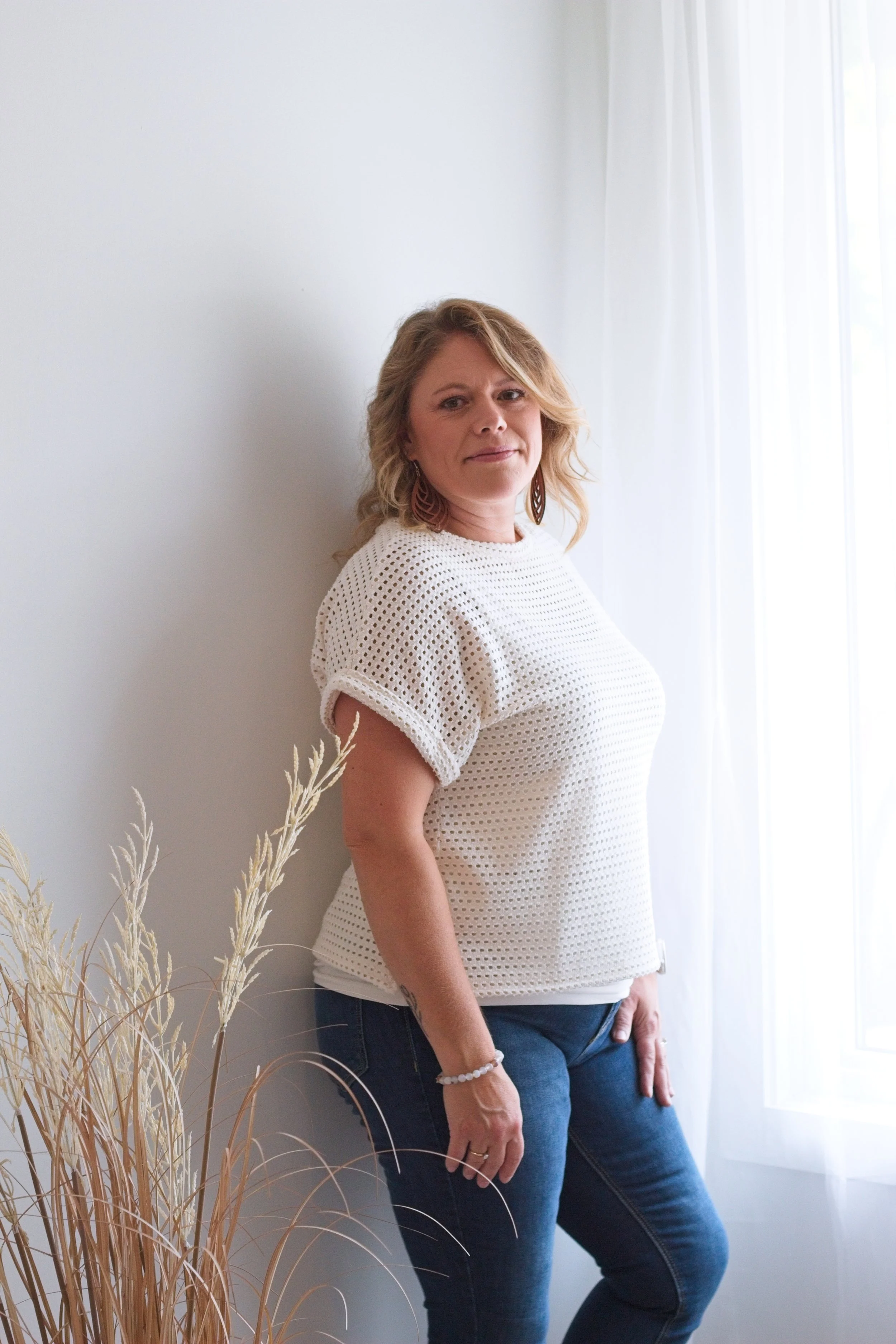 A woman with light brown, curly hair standing by a white wall and sheer white curtains, wearing a cream crochet top and blue jeans, looking at the camera.