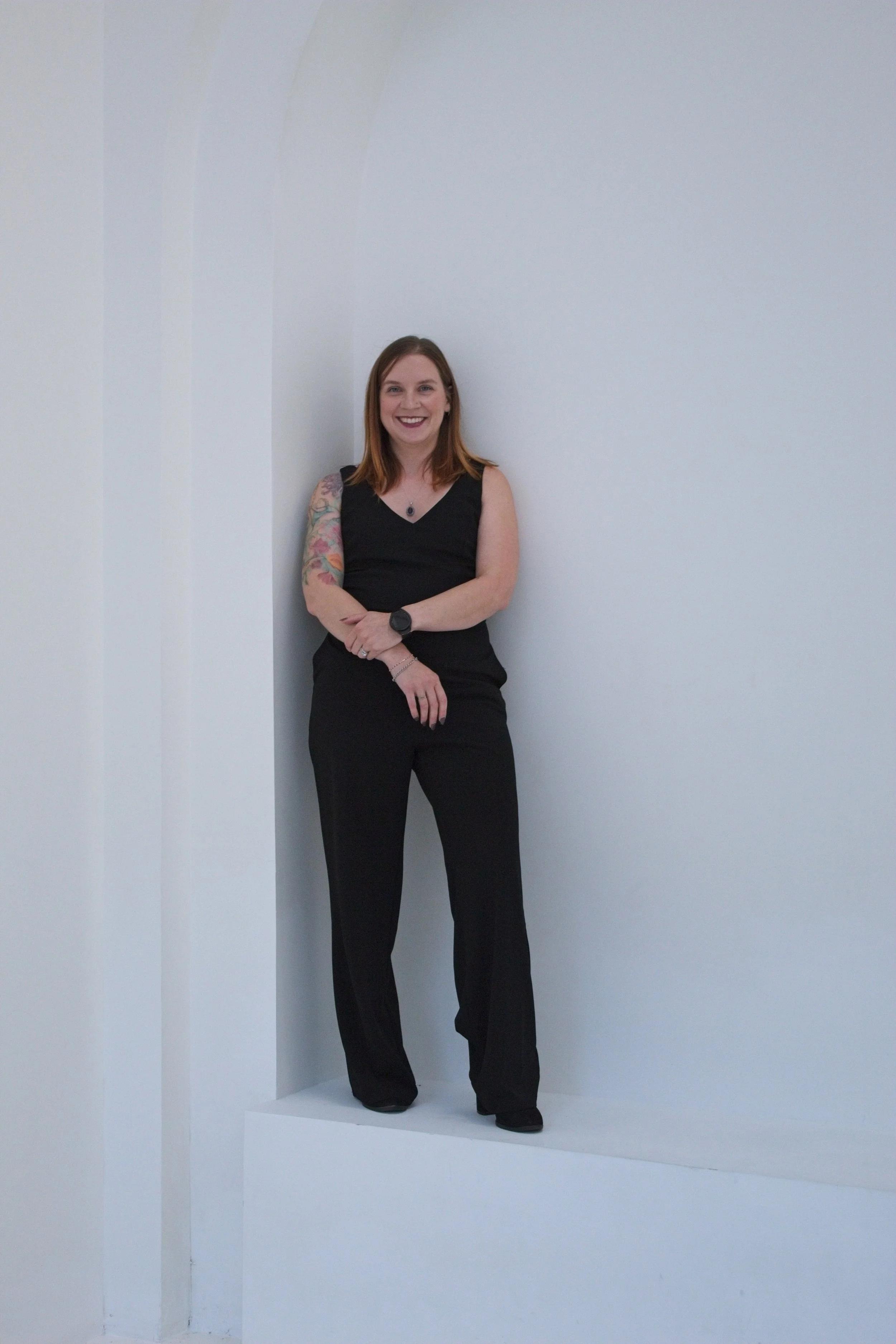 A woman with shoulder-length red hair, smiling, wearing a black sleeveless top and black pants, standing with arms crossed in a white, minimalist, arched corner space.