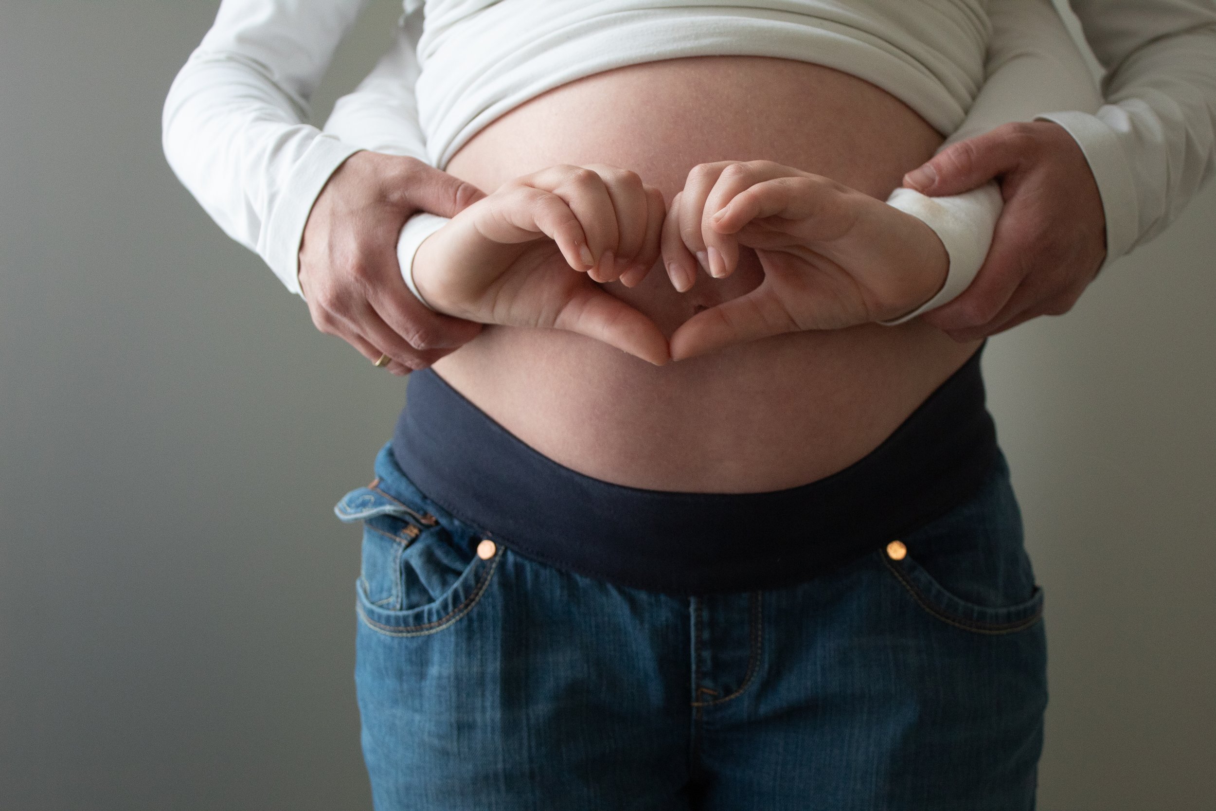 A pregnant woman making a heart shape with her hands over her belly, with a person's hands supporting hers.