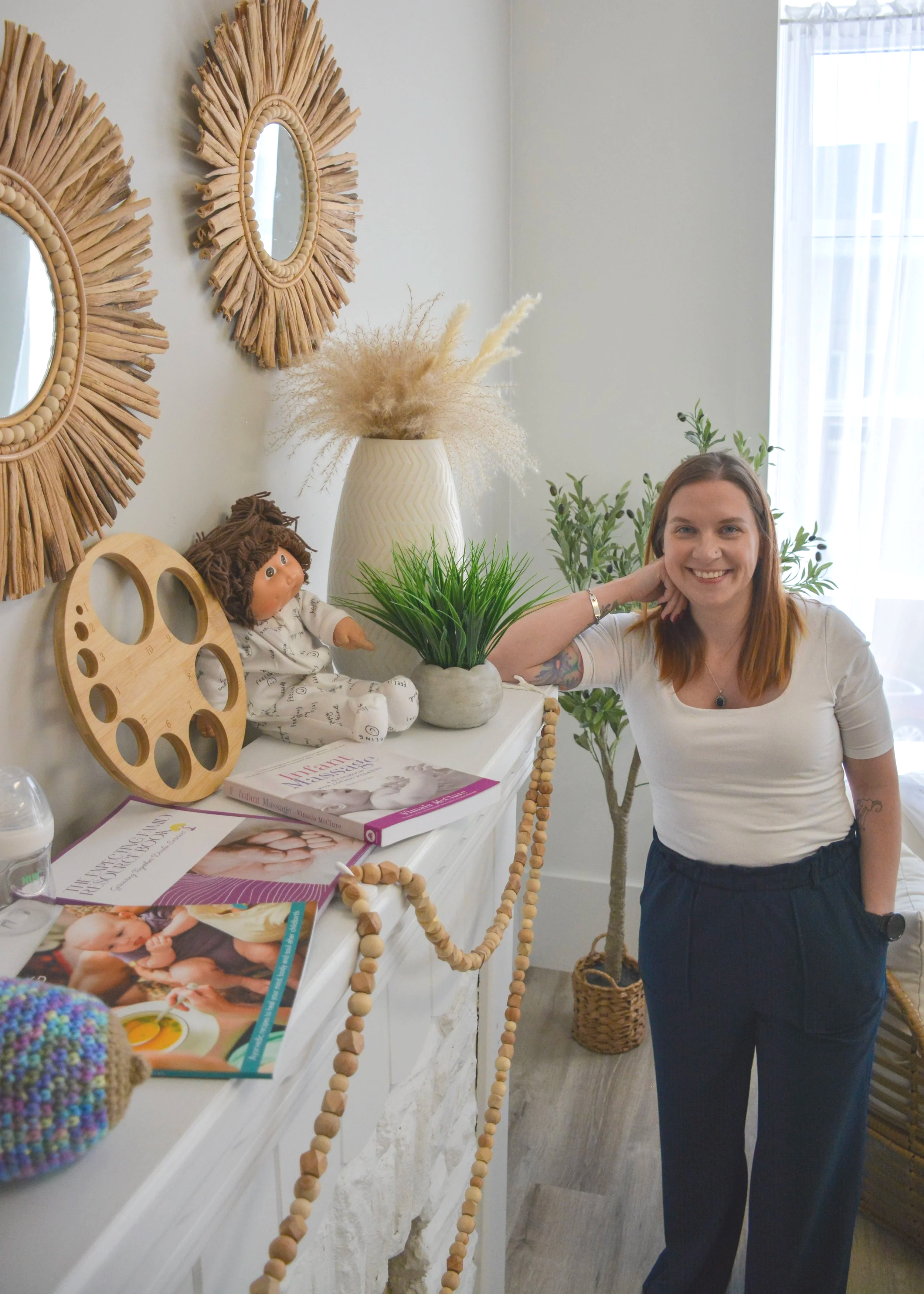 A woman standing by a white dresser in a well-lit room, smiling. There are several decorative items on the dresser including a doll, a large green plant, a white vase with pampas grass, and magazines. The wall behind has two round, straw-framed mirrors. There is a potted plant and a window with sheer curtains in the background.
