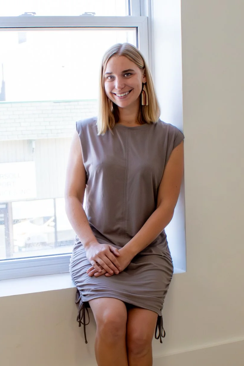 A young woman with shoulder-length blonde hair, smiling and sitting on a windowsill in front of a large window, wearing a sleeveless taupe dress and matching earrings.