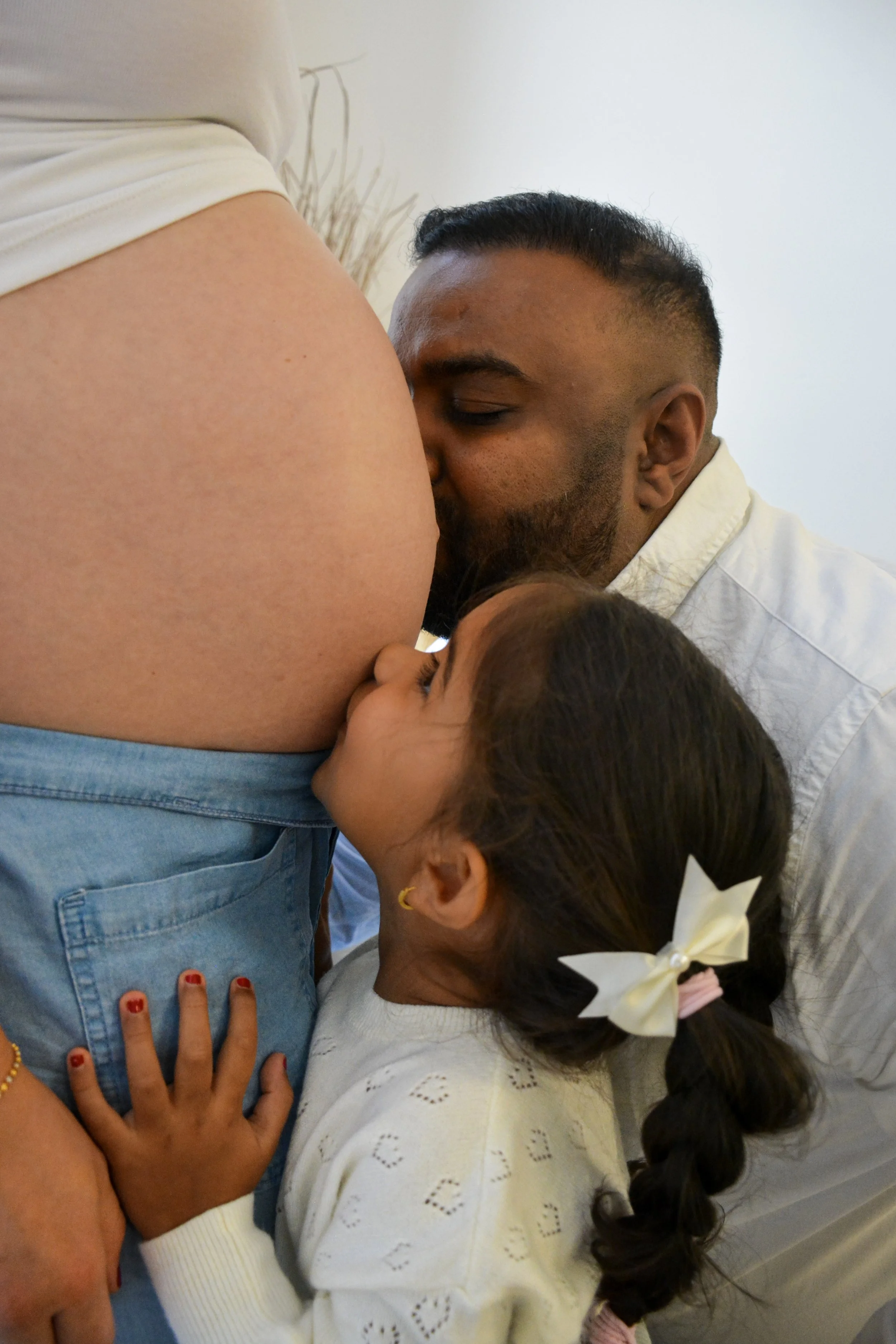 A family moment where a pregnant woman is being kissed on her belly by a man while a young girl is kissing her mother’s belly, all around her pregnant stomach.