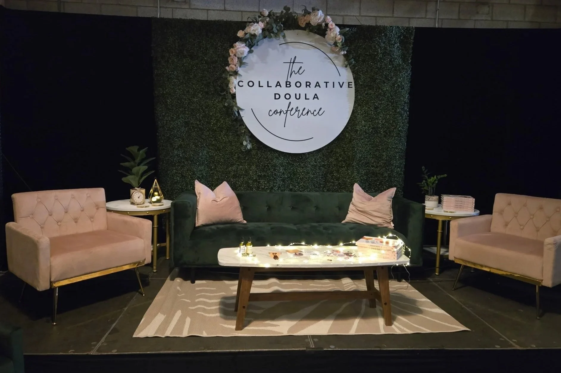 Conference stage setup with a green couch, two pink armchairs, and a large round sign that reads 'The Collaborative Doula Conference' in London. Ontario.